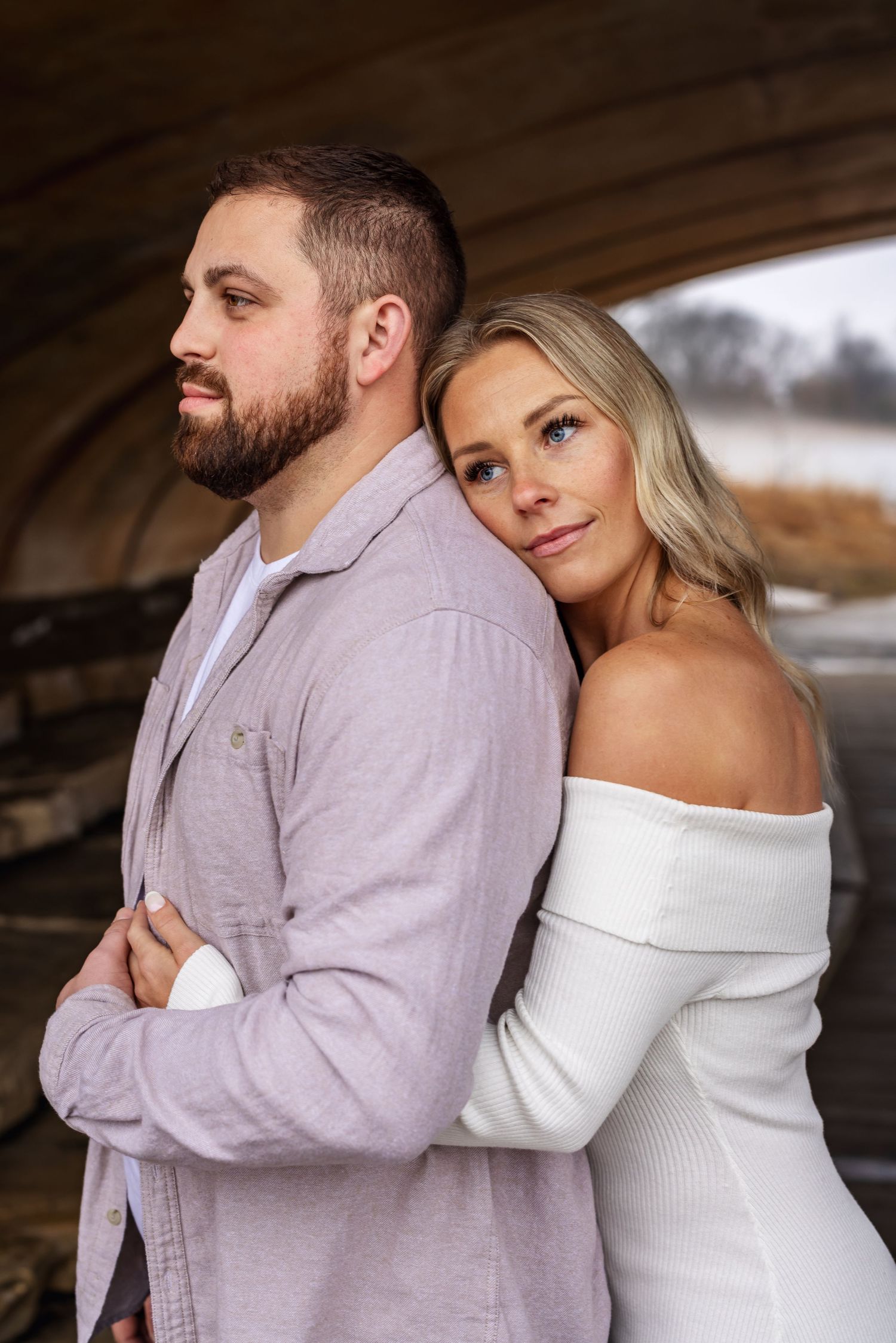 A romantic couple poses together in soft lighting under an arch, wearing coordinating lavender and white outfits.