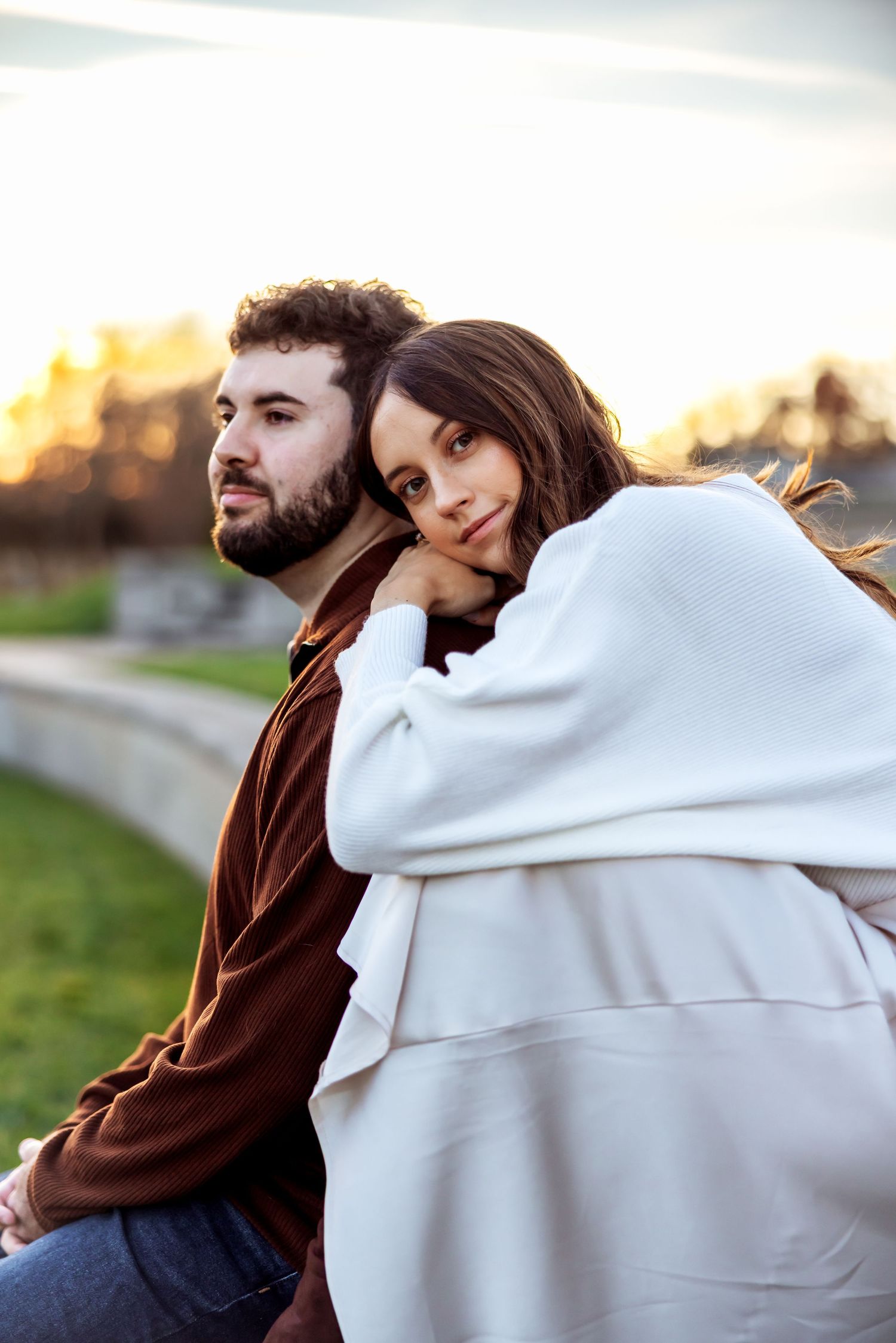A couple sits close together outdoors at sunset, with one wearing a brown sweater and the other in cozy white layers.