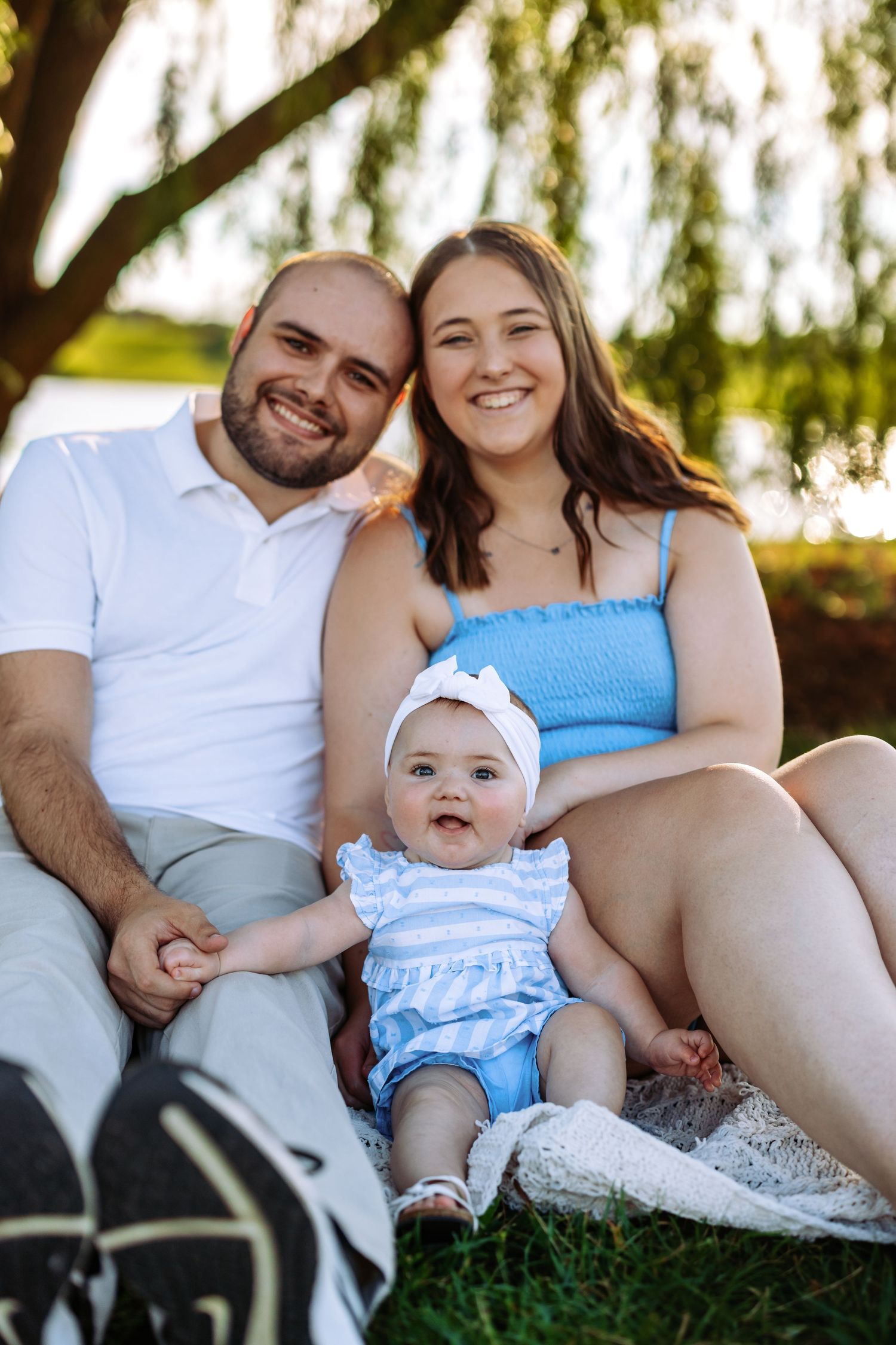Family sits together in grass against willow tree backdrop during outdoor photo session at sunset.