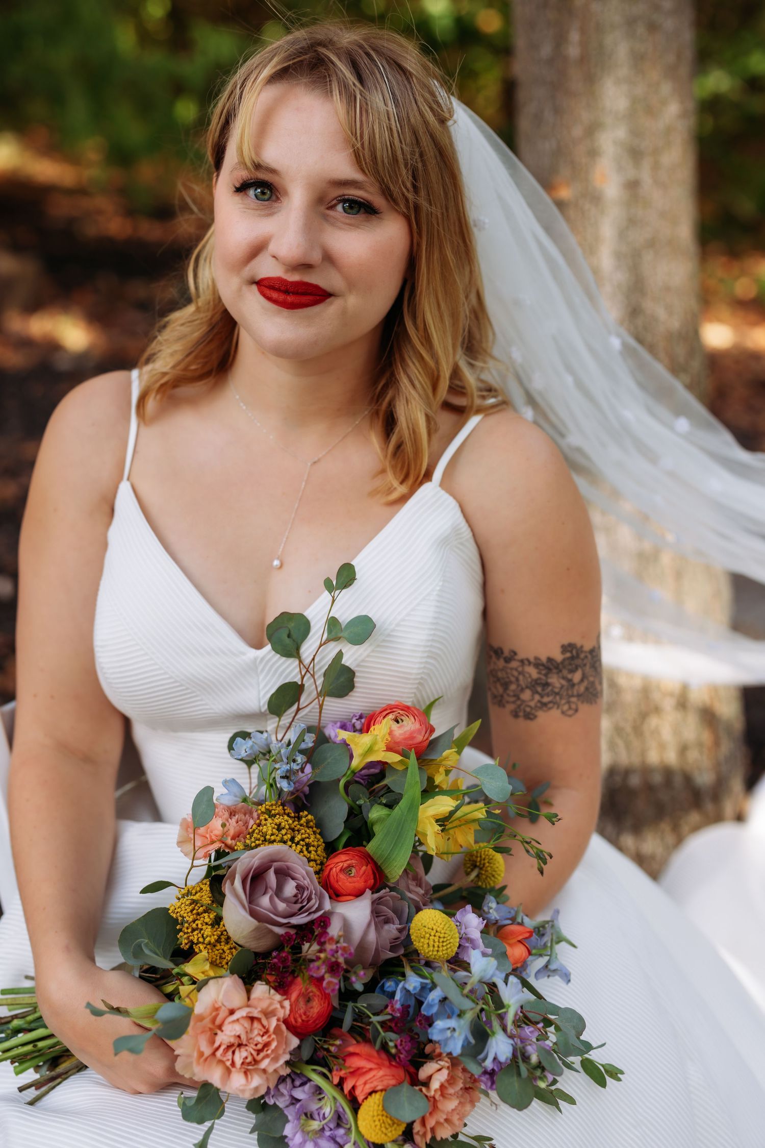 A bride in a white dress holds a vibrant rainbow-colored wedding bouquet outdoors.