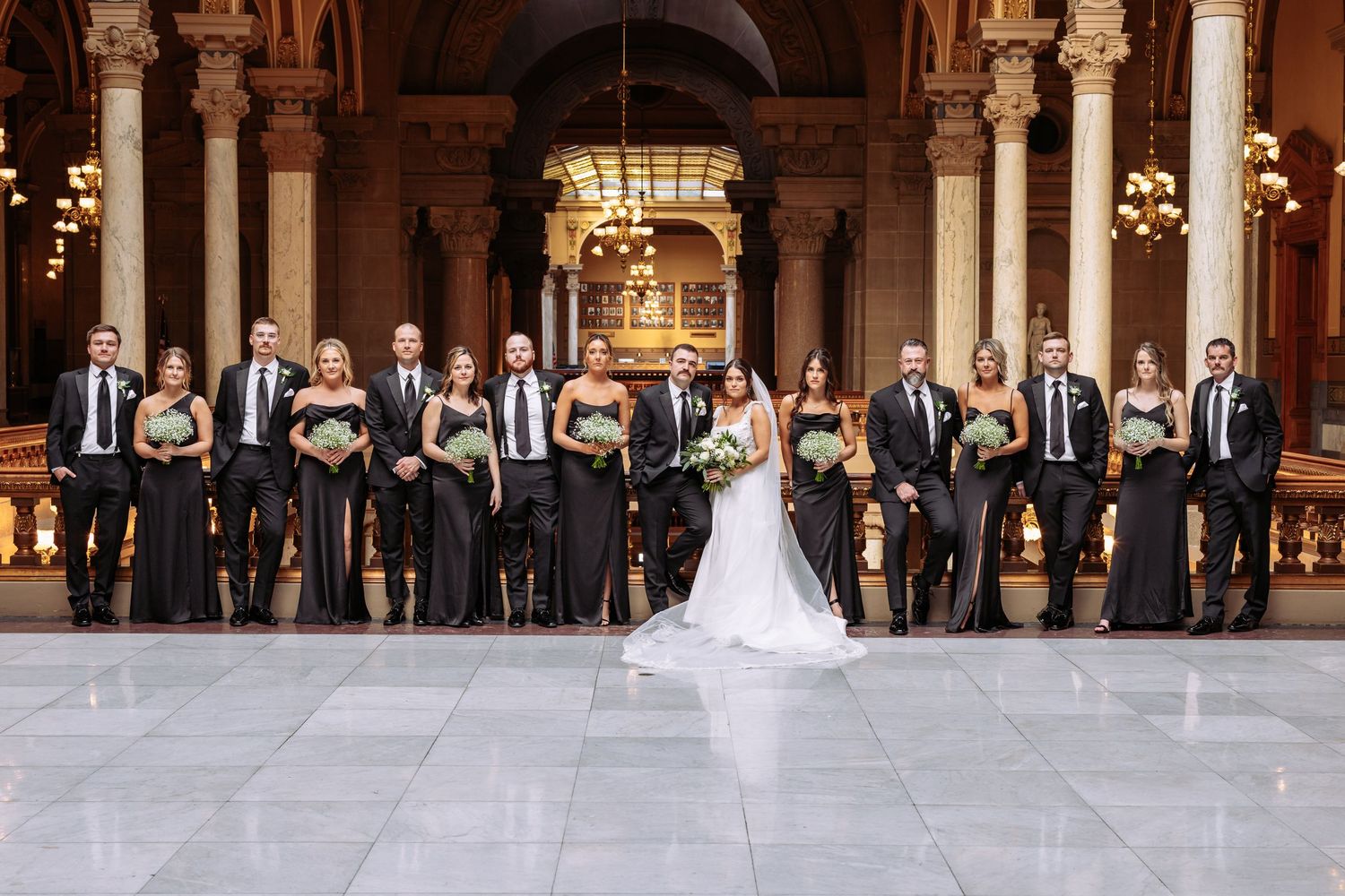 Wedding party in formal black attire poses elegantly in a grand columned hall with ornate architecture.