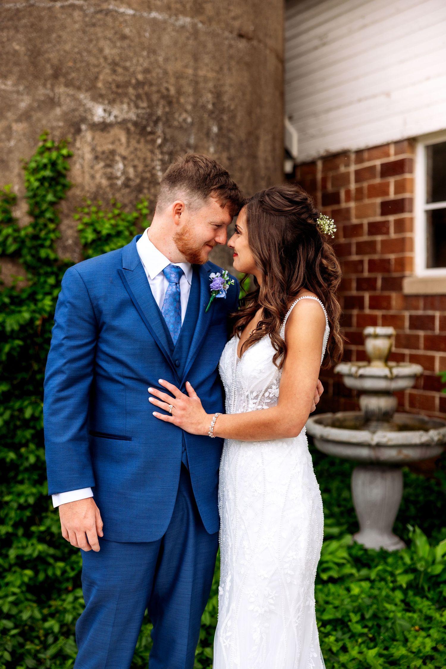 A bride and groom share a romantic moment outdoors, she in a white lace gown, he in a blue suit with floral boutonniere.
