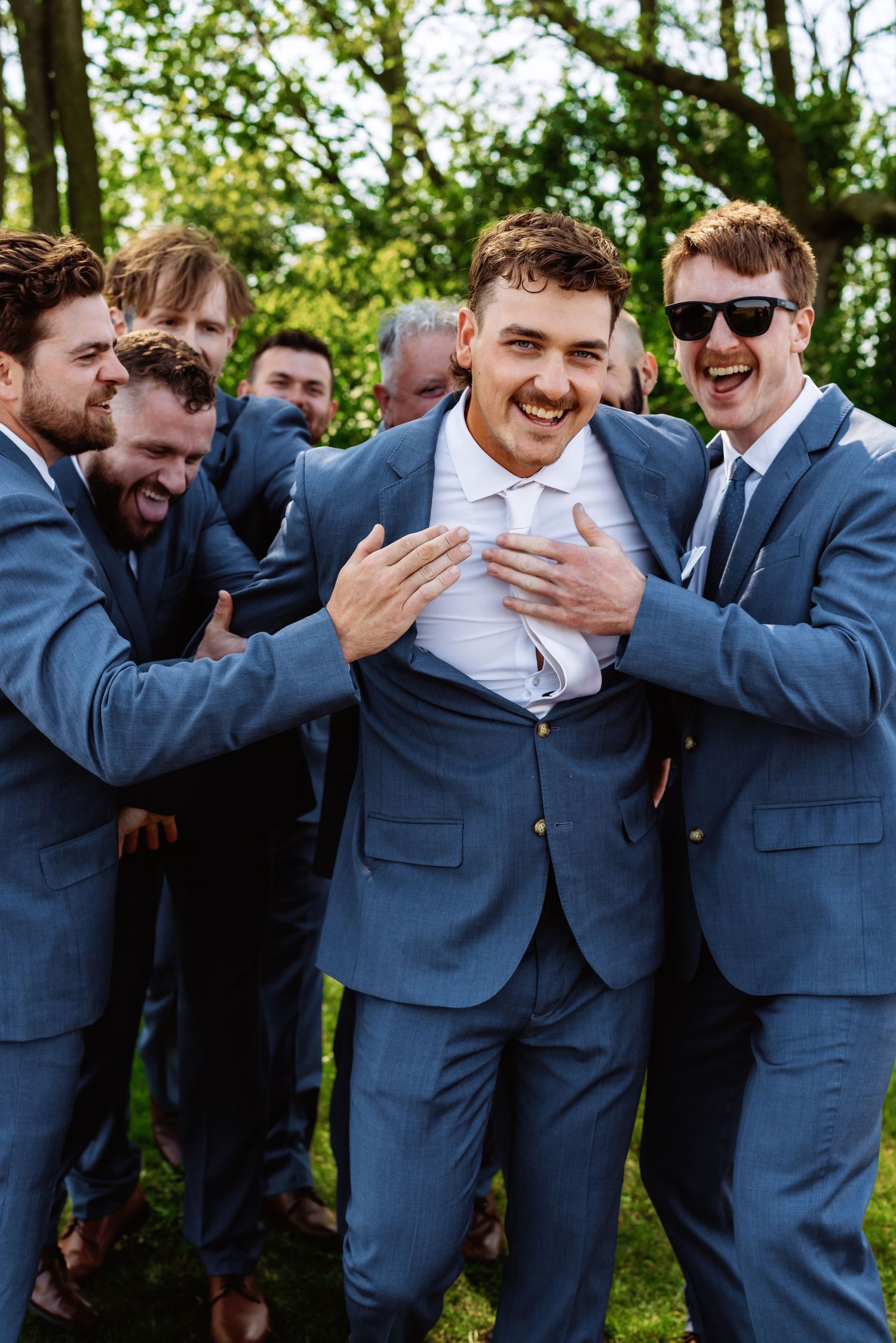 A group of groomsmen in matching blue suits share a candid laughing moment together outdoors.