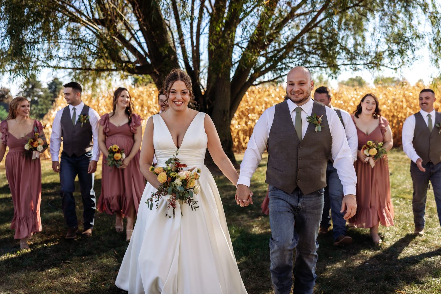 Wedding party stands together in formal attire on a sunny autumn day with trees in the background.