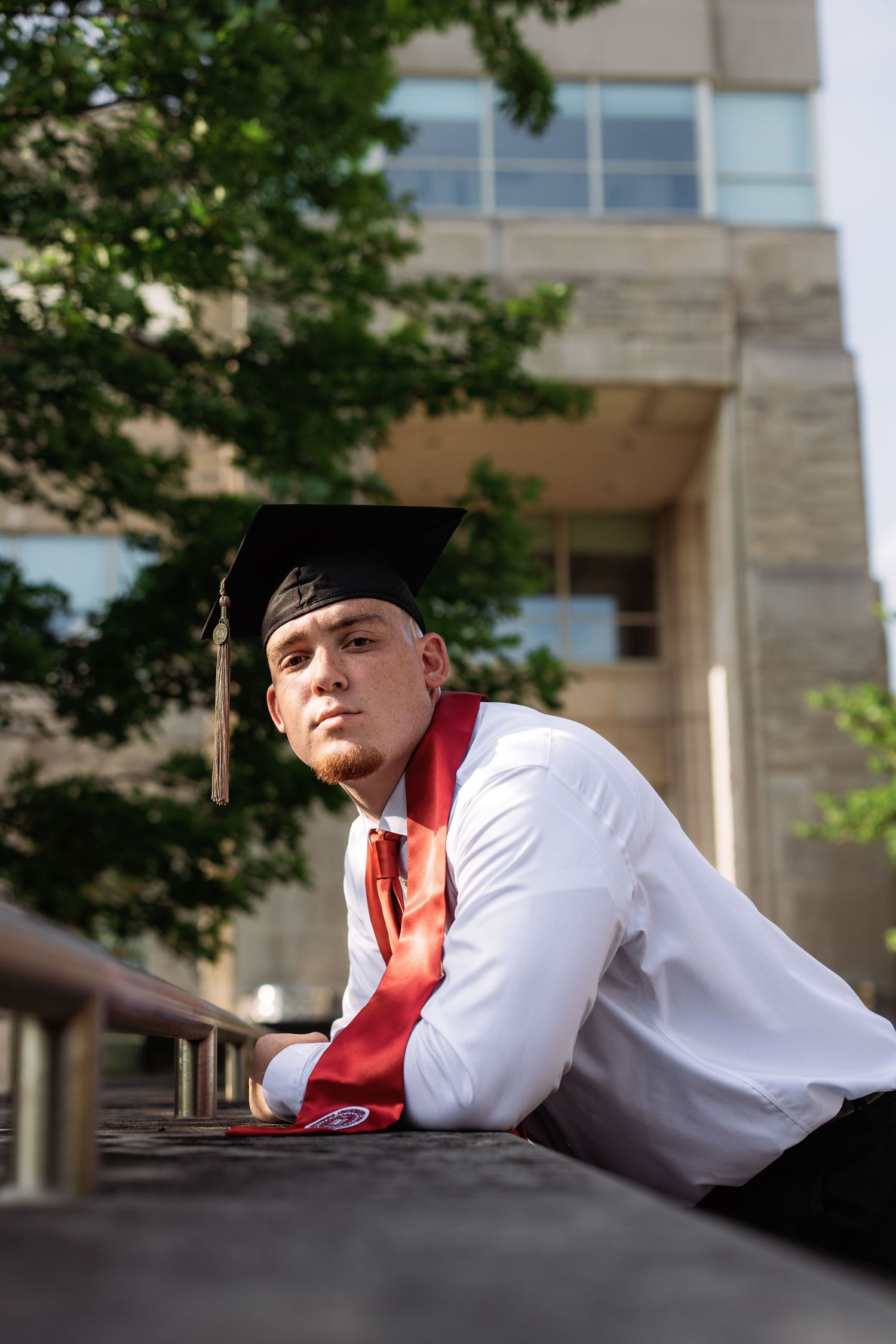 Graduate in cap and gown lying on bench in contemplative poses outside university building.