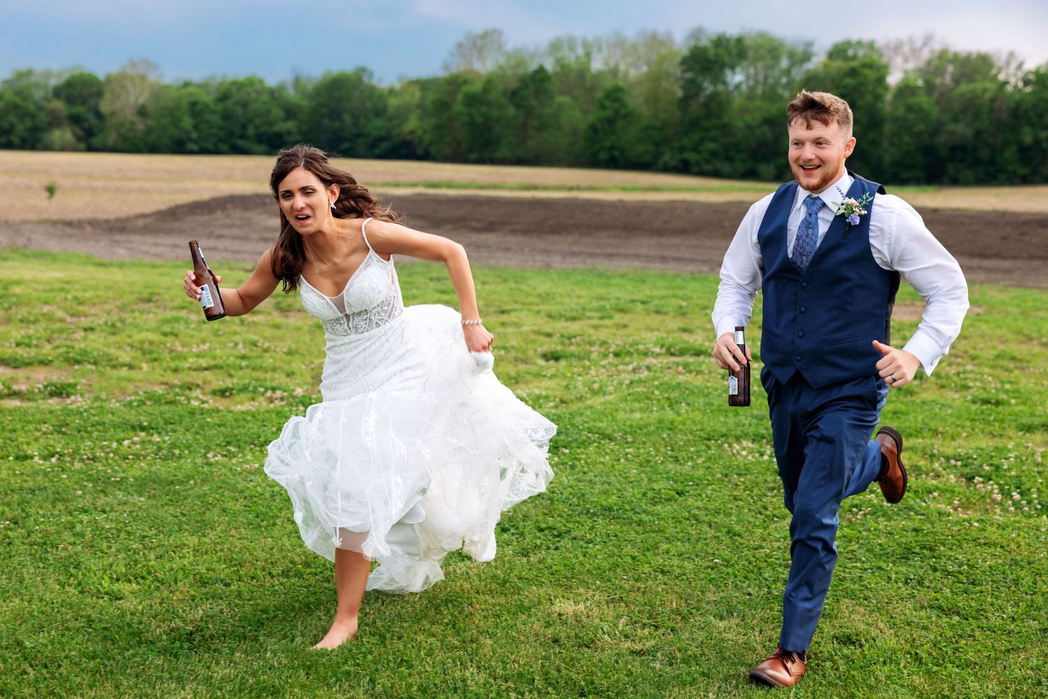 Joyful bride and groom running barefoot through green field holding beer bottles on their wedding day.