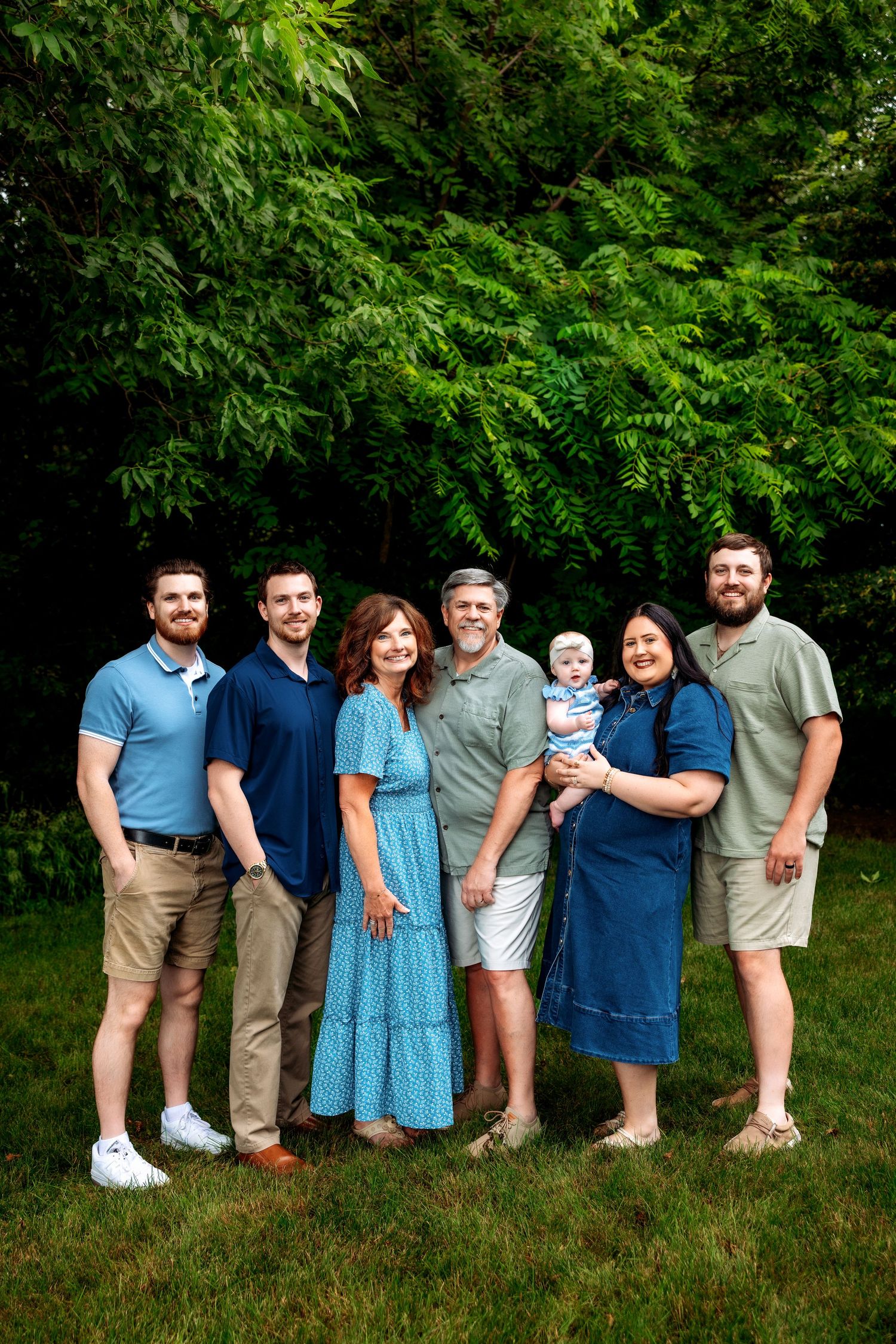 Multi-generational family of seven smiling outdoors on green lawn with lush trees, including baby held by young woman in blue dress.