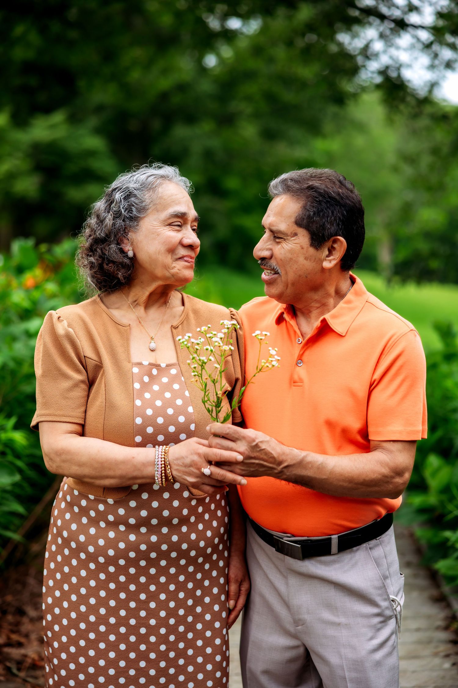 Elderly couple smiling at each other in a lush garden, holding small yellow wildflowers, wearing casual brown and orange attire.