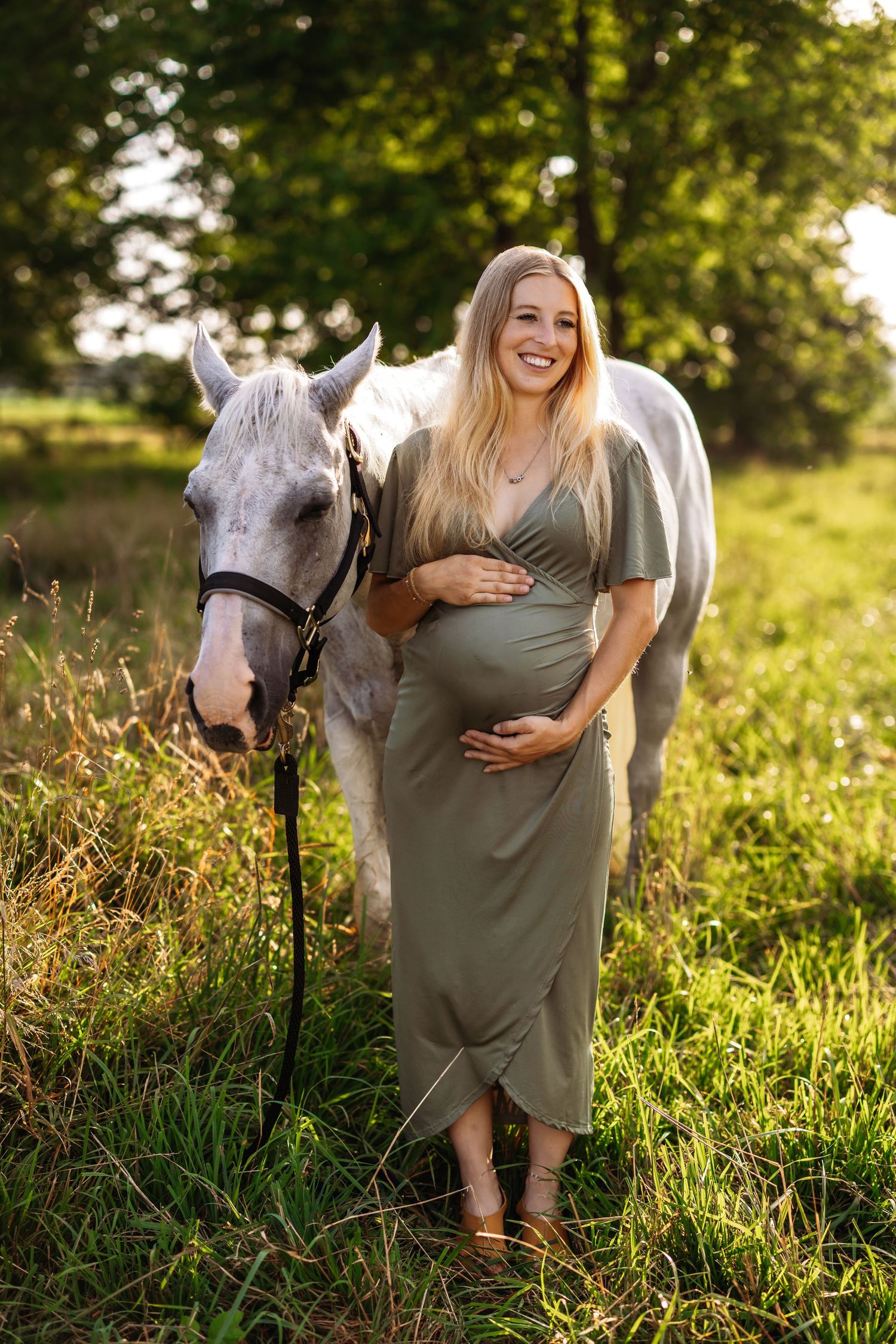 An expectant woman in a gray dress stands with a white horse in a sunlit field during golden hour.