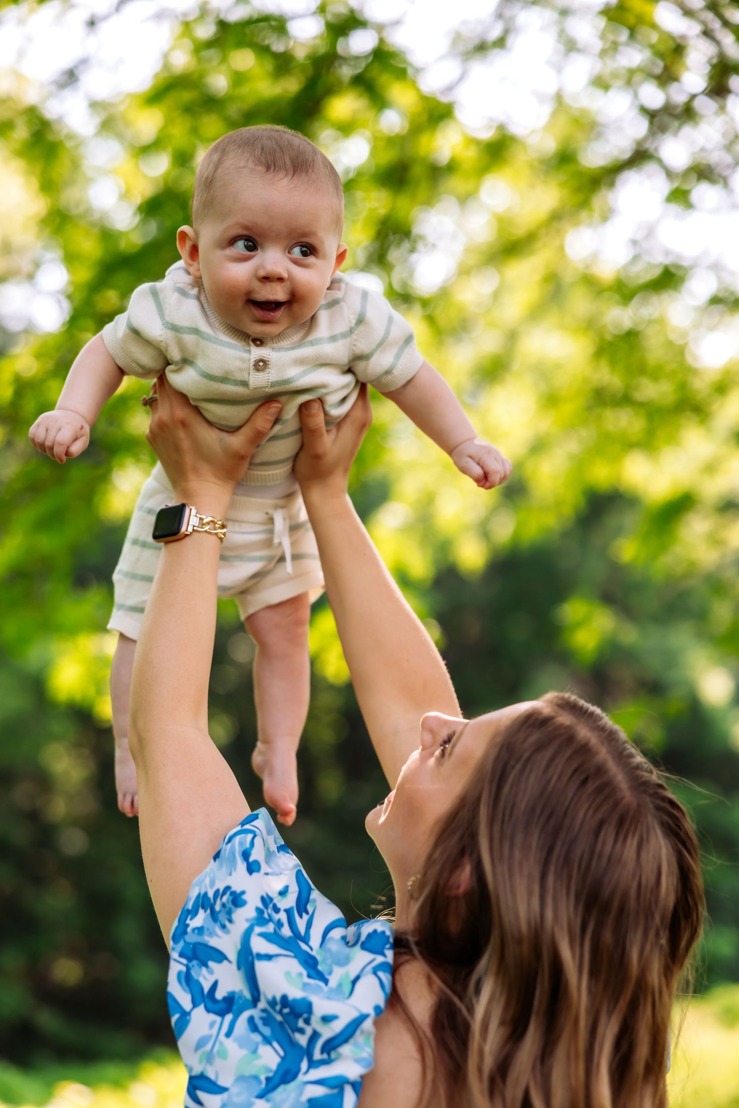 A baby in a beige outfit is lifted high in the air against a sunlit green background at the park.