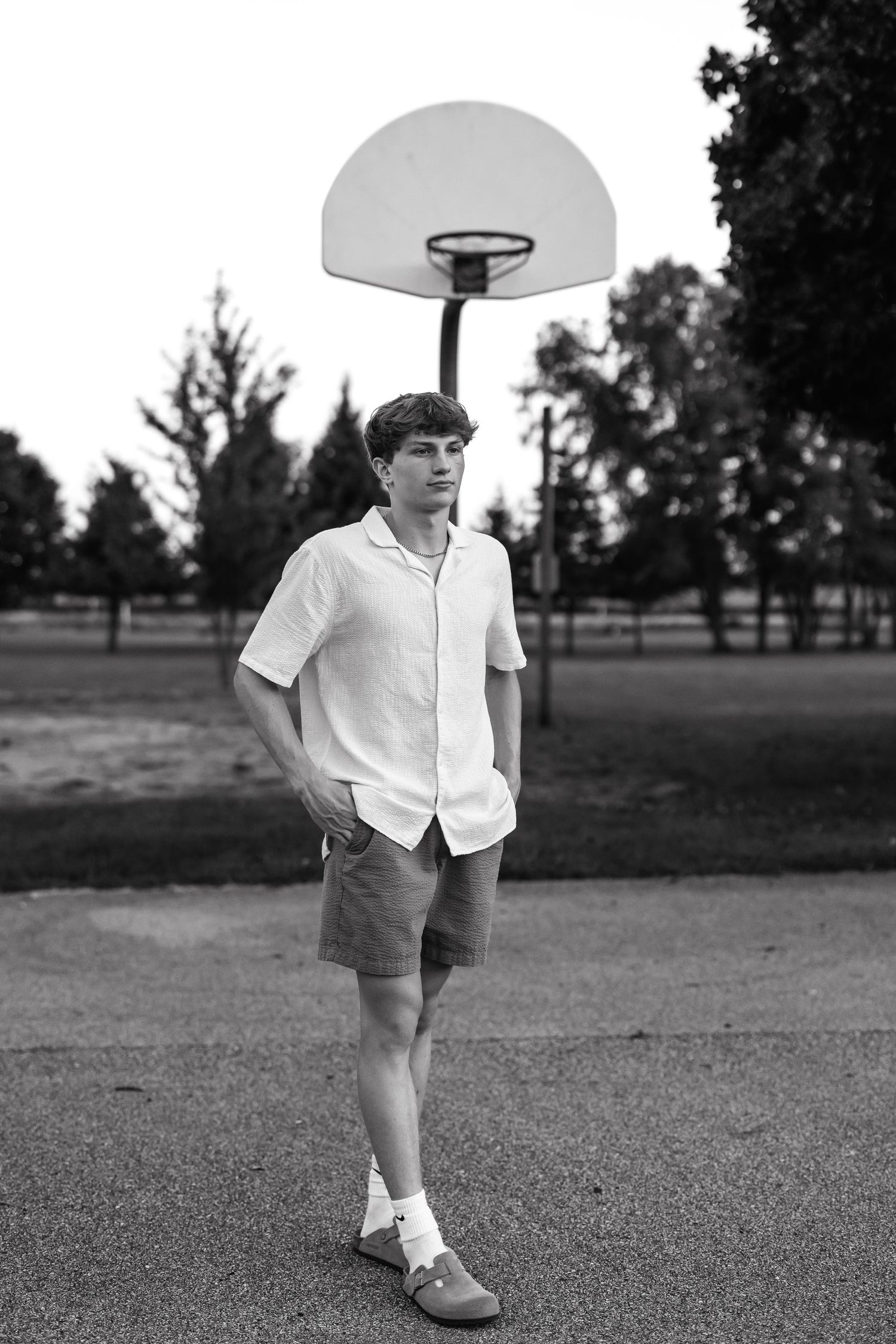 Young person in casual attire stands near a basketball hoop on an outdoor court.