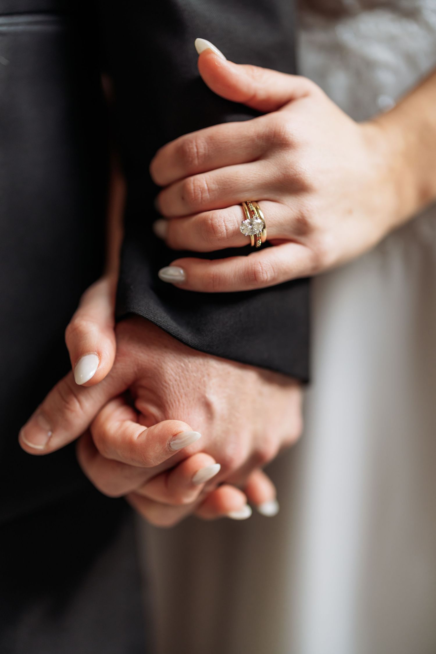Close-up of intertwined hands with engagement ring and neutral manicured nails against dark fabric background.