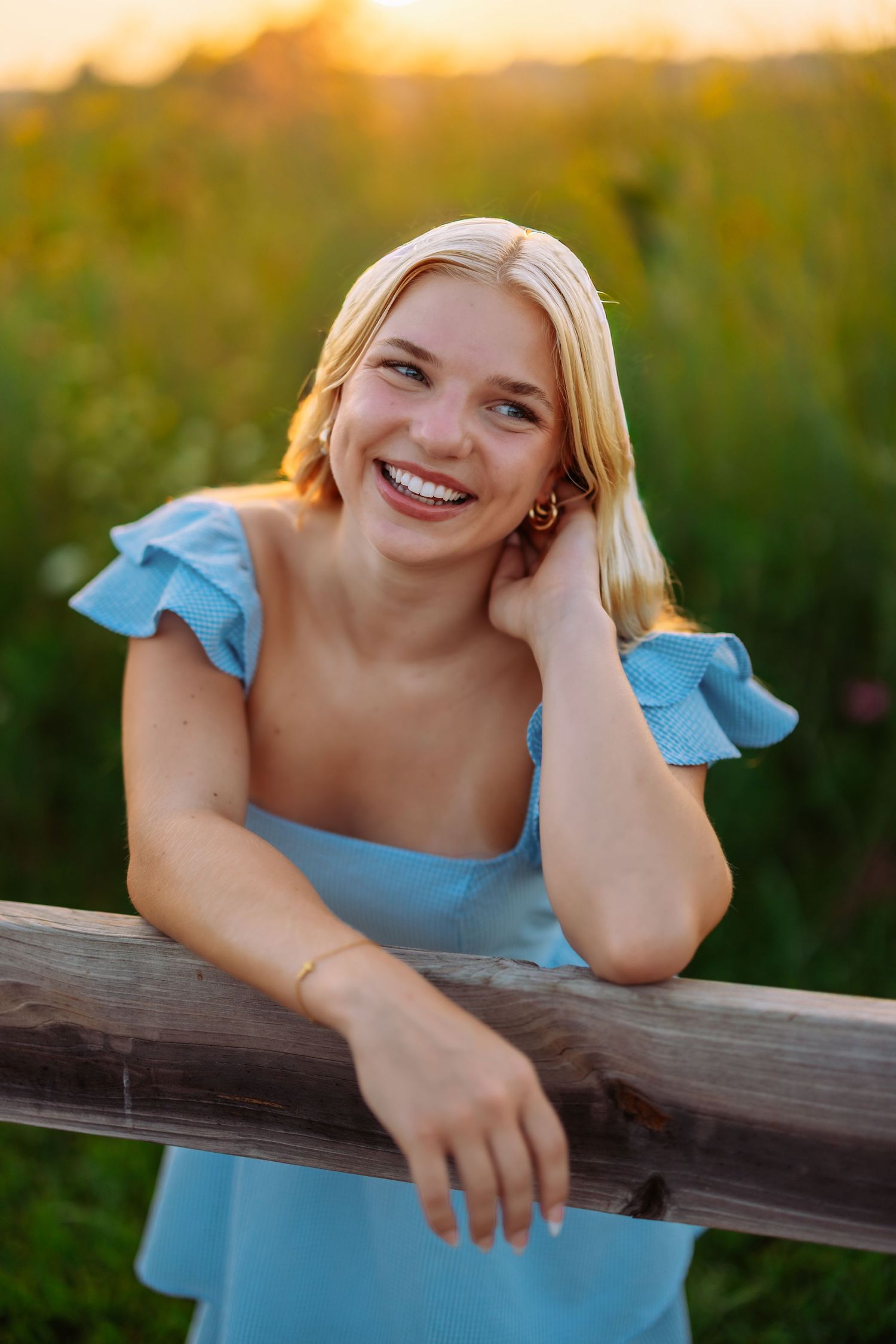 A joyful portrait in a light blue dress leaning on a wooden fence during golden hour sunset.