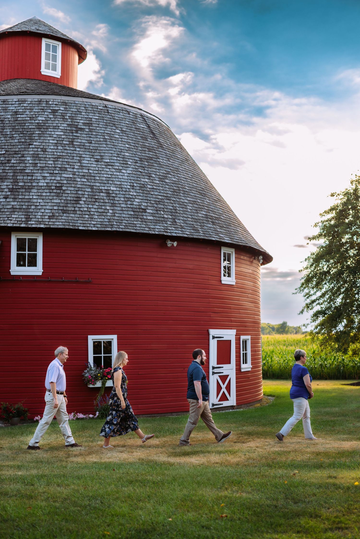 People walk in a line around a large red round barn in a rural setting during golden hour.