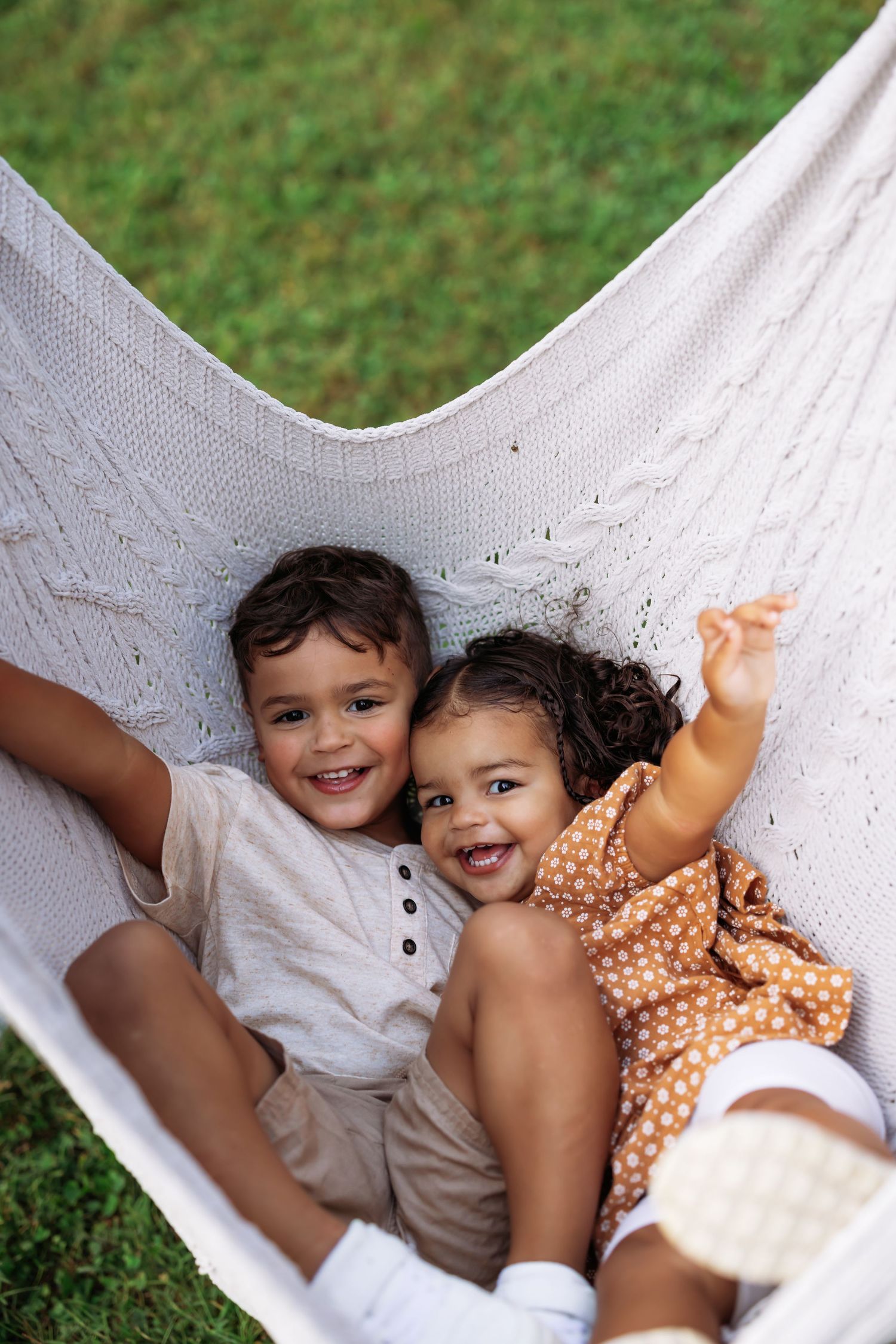 Two children laying together in a white hammock outdoors on grass, smiling and playing.
