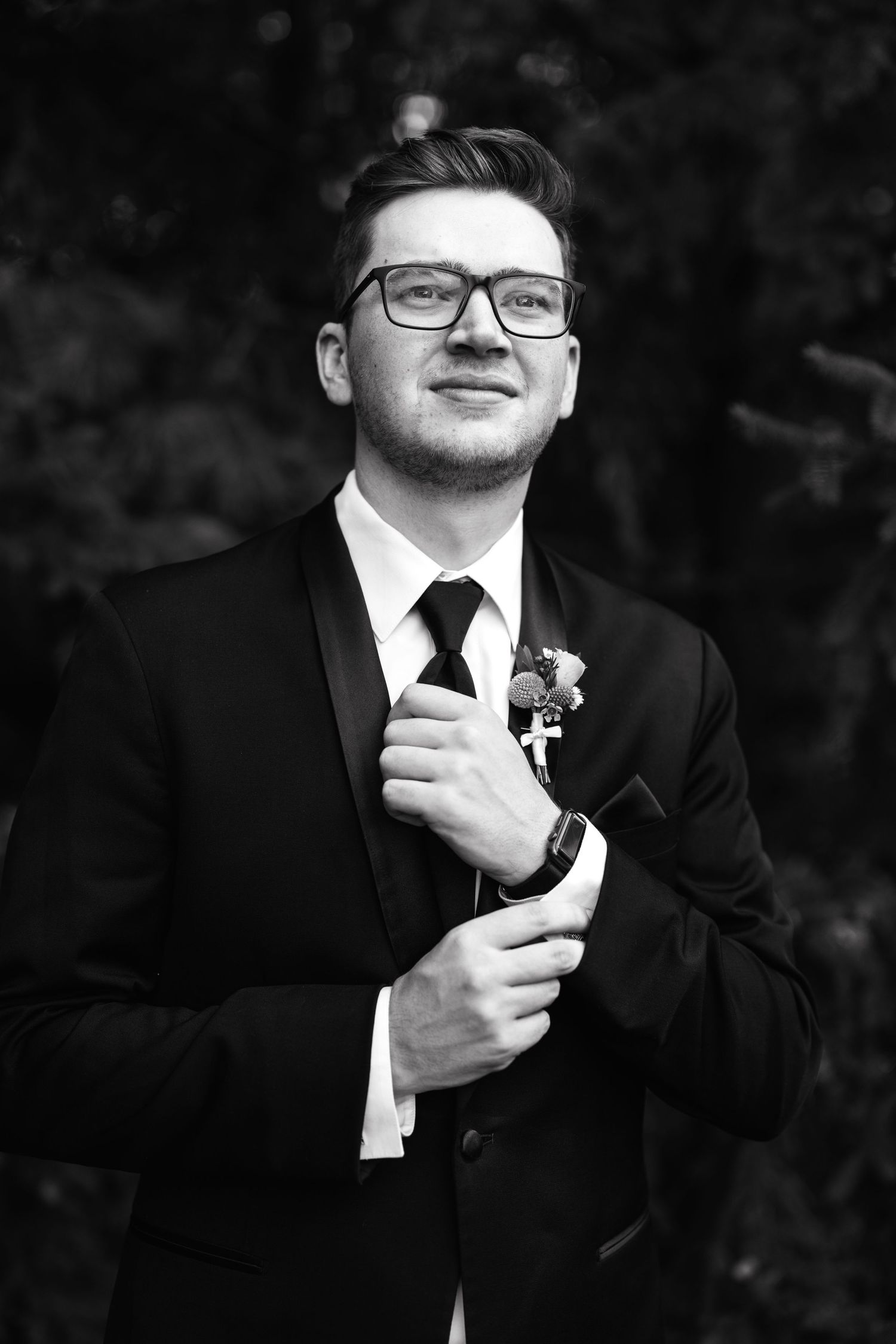 Professional portrait of a groom in black suit and glasses adjusting his tie during a wedding photoshoot.
