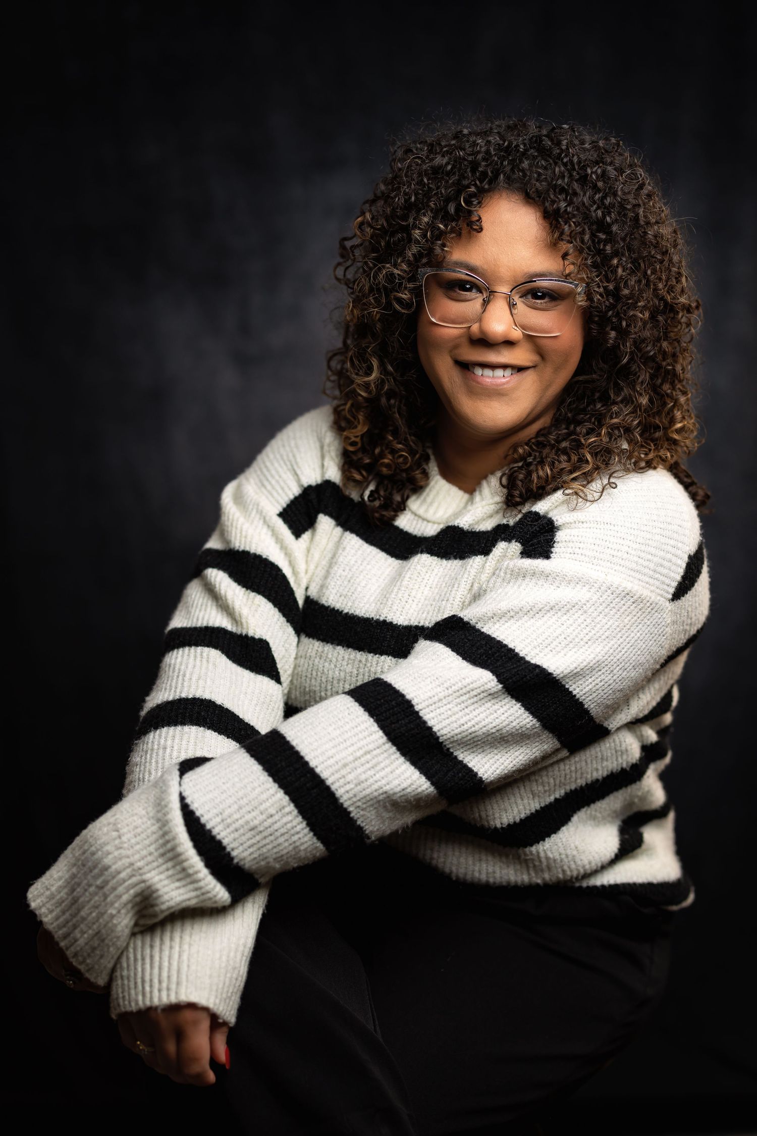 Portrait of person wearing striped sweater with curly hair and glasses against dark background.