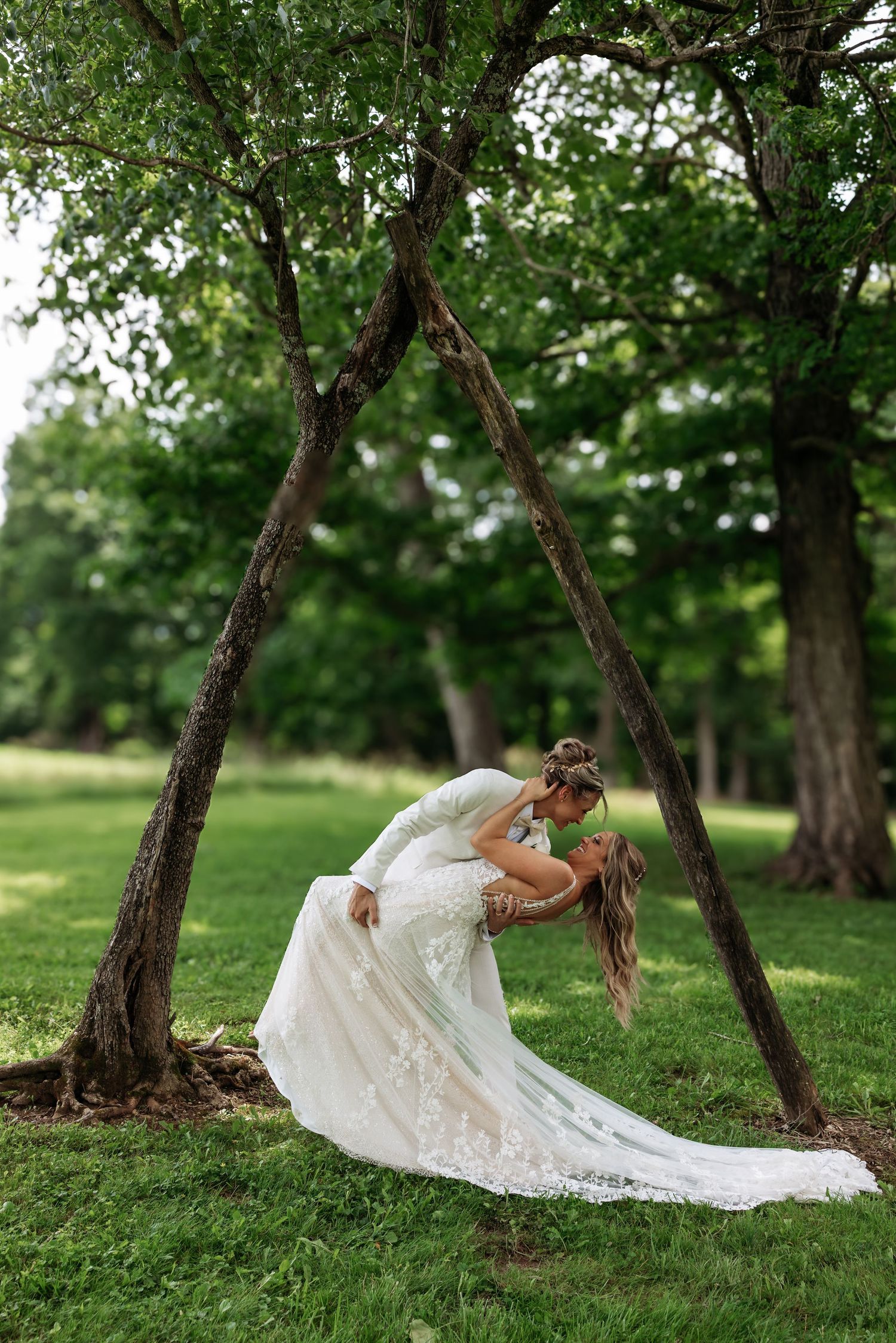 Bride in flowing white lace gown lies gracefully on grass under trees in rustic outdoor setting.