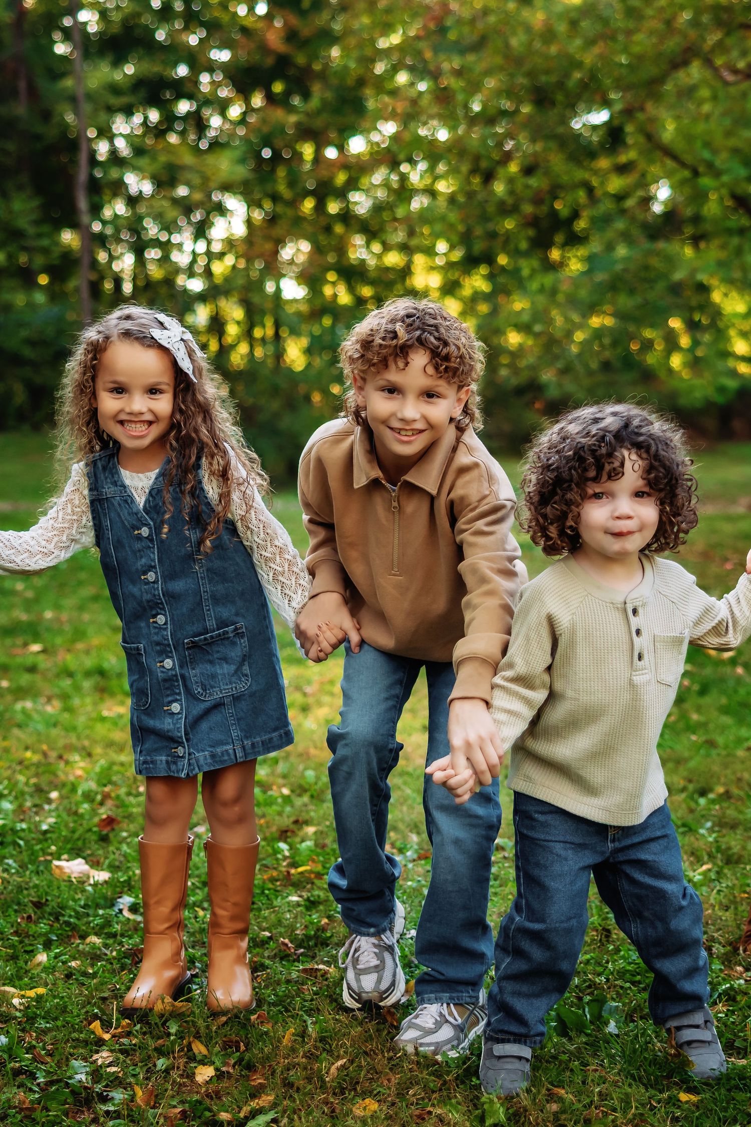 Three children hold hands and walk together through a grassy field on an autumn afternoon.