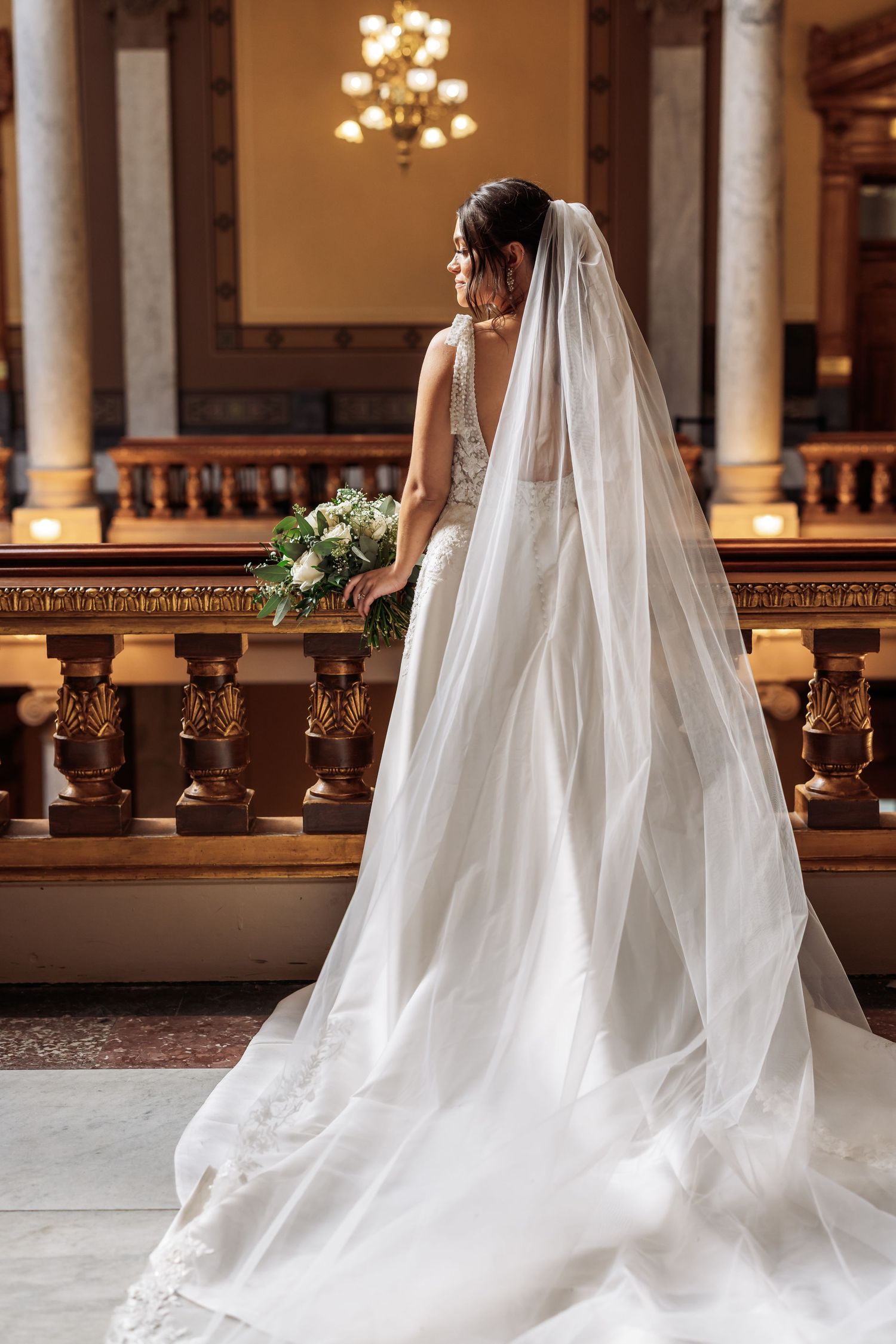 A bride in an elegant white gown with a flowing cathedral veil stands in a historic church holding a bouquet.