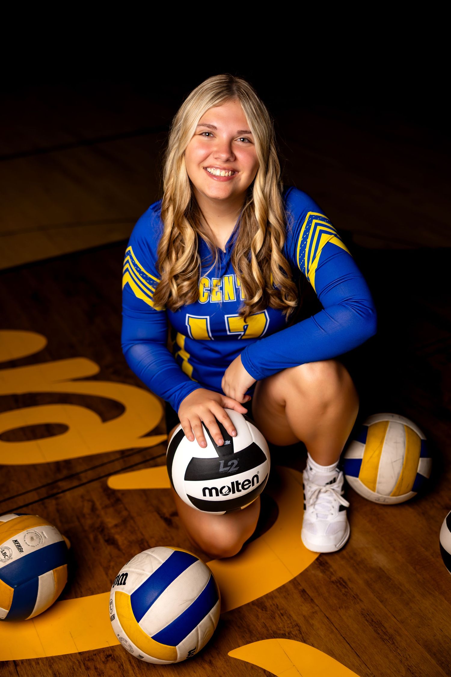 Smiling female volleyball player in blue and yellow uniform posing with Molten volleyballs on a gym floor.