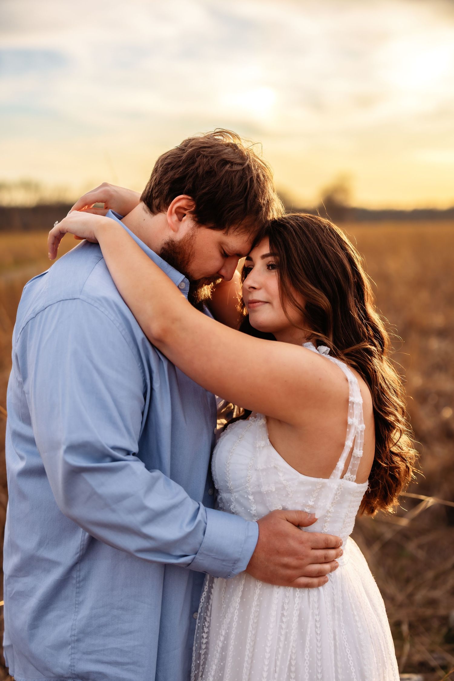 A romantic couple embraces in a field at sunset, with warm golden light surrounding them in their formal attire.