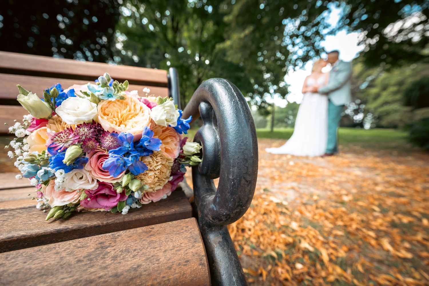 Colorful wedding bouquet on a park bench with bride and groom blurred in the background among autumn leaves.