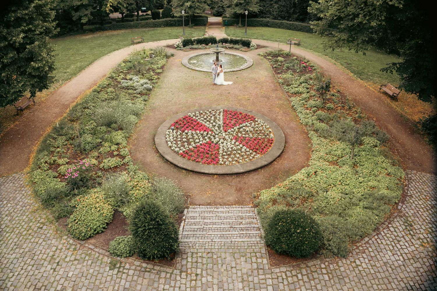 Aerial view of a formal garden with geometric floral patterns, brick pathways, and a central fountain surrounded by lush greenery.