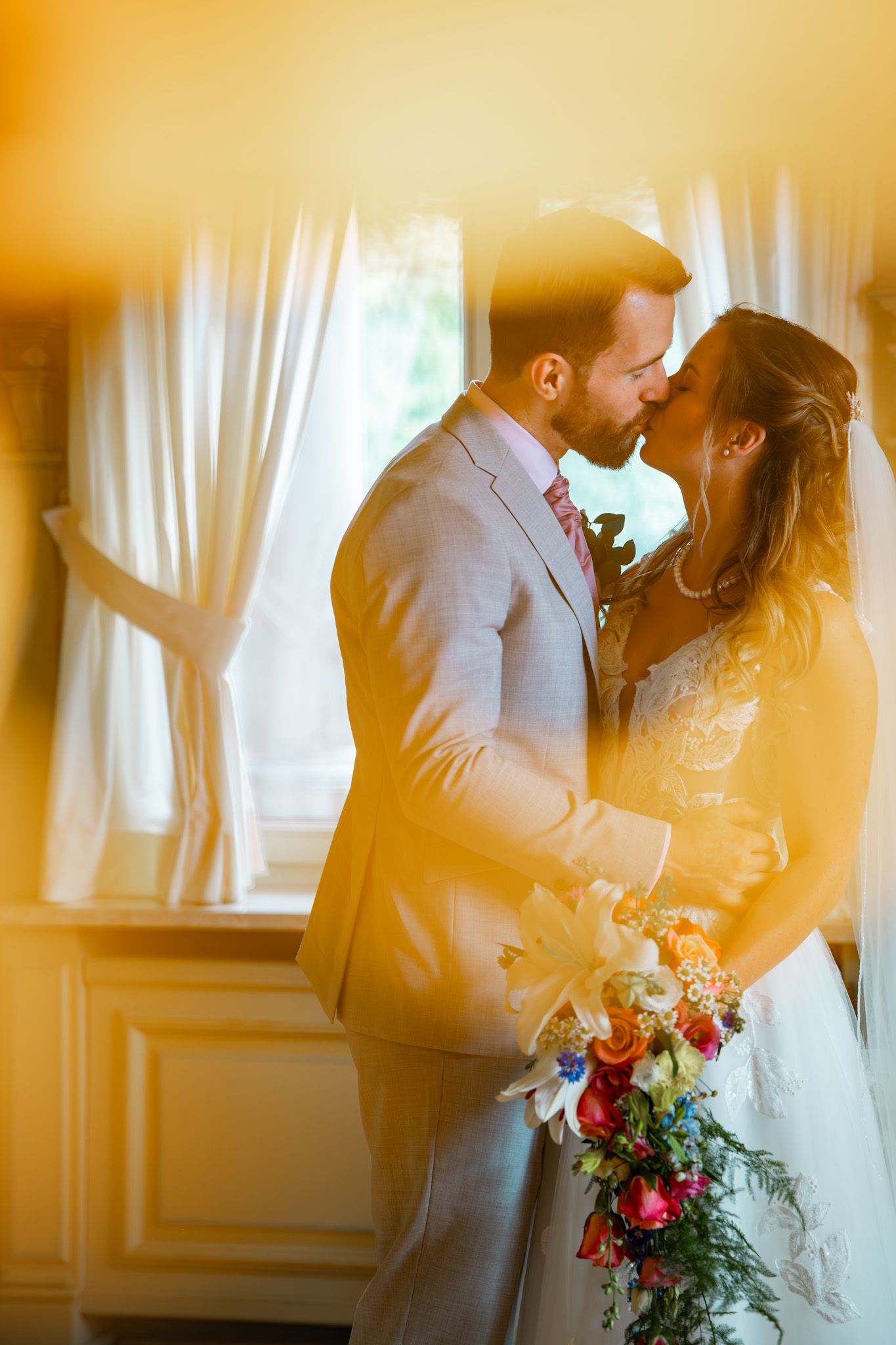 Bride and groom share a romantic kiss indoors, bride holding a colorful bouquet, bathed in warm golden light.