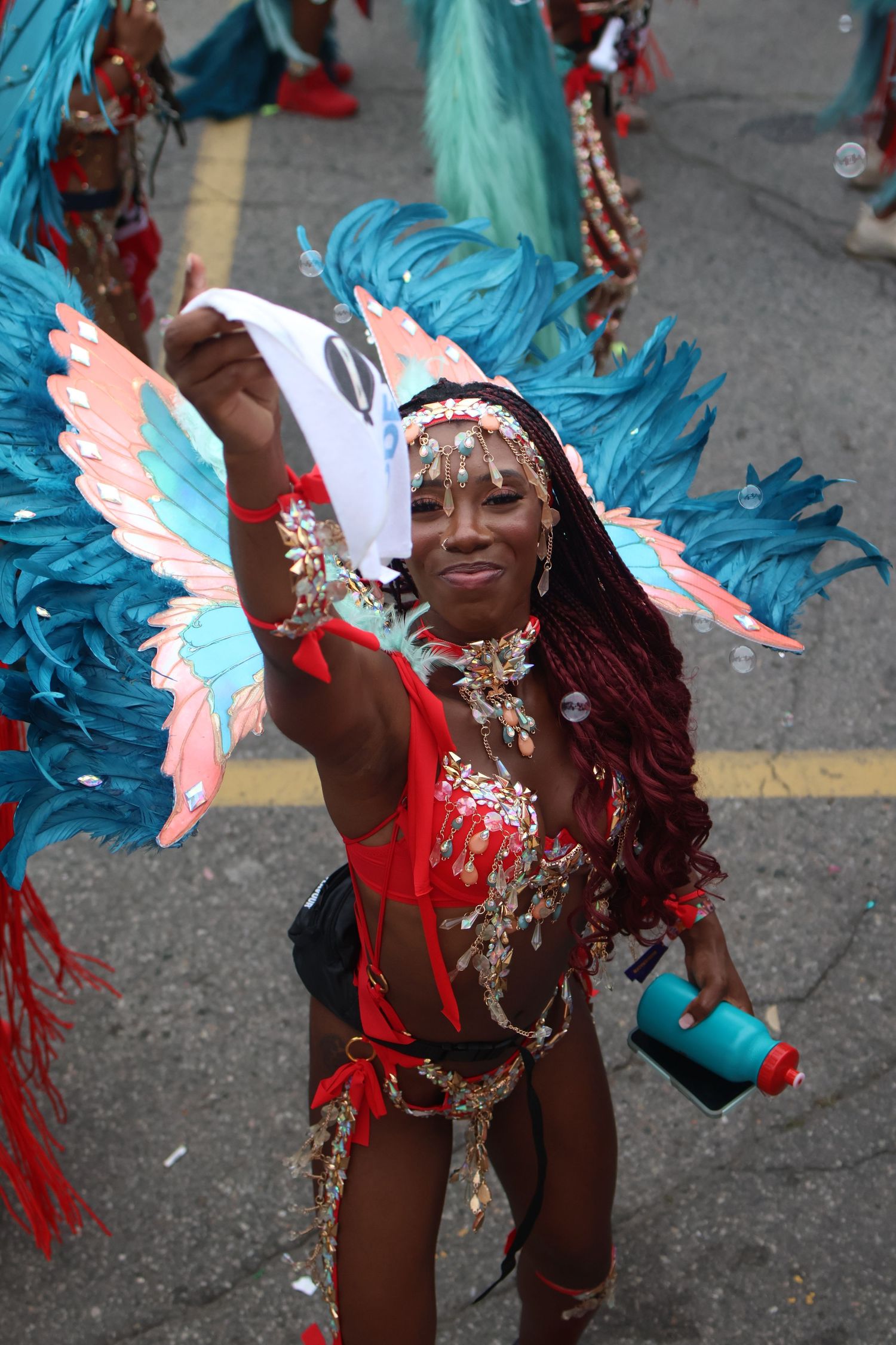 Carnival performer in bright red costume with turquoise feathered wings dances energetically during street parade.