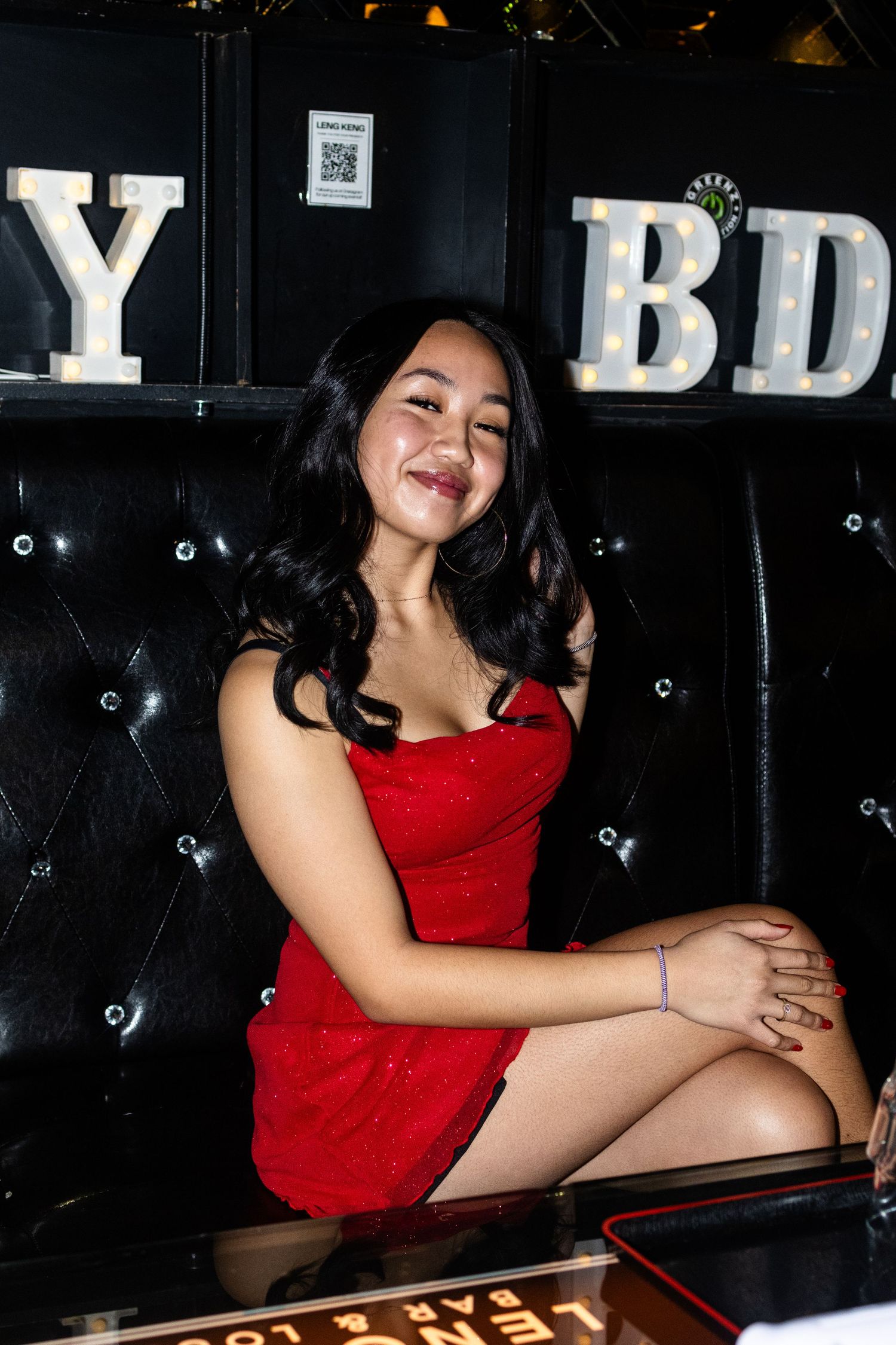 Woman in red dress poses on black leather couch with illuminated letters in background.