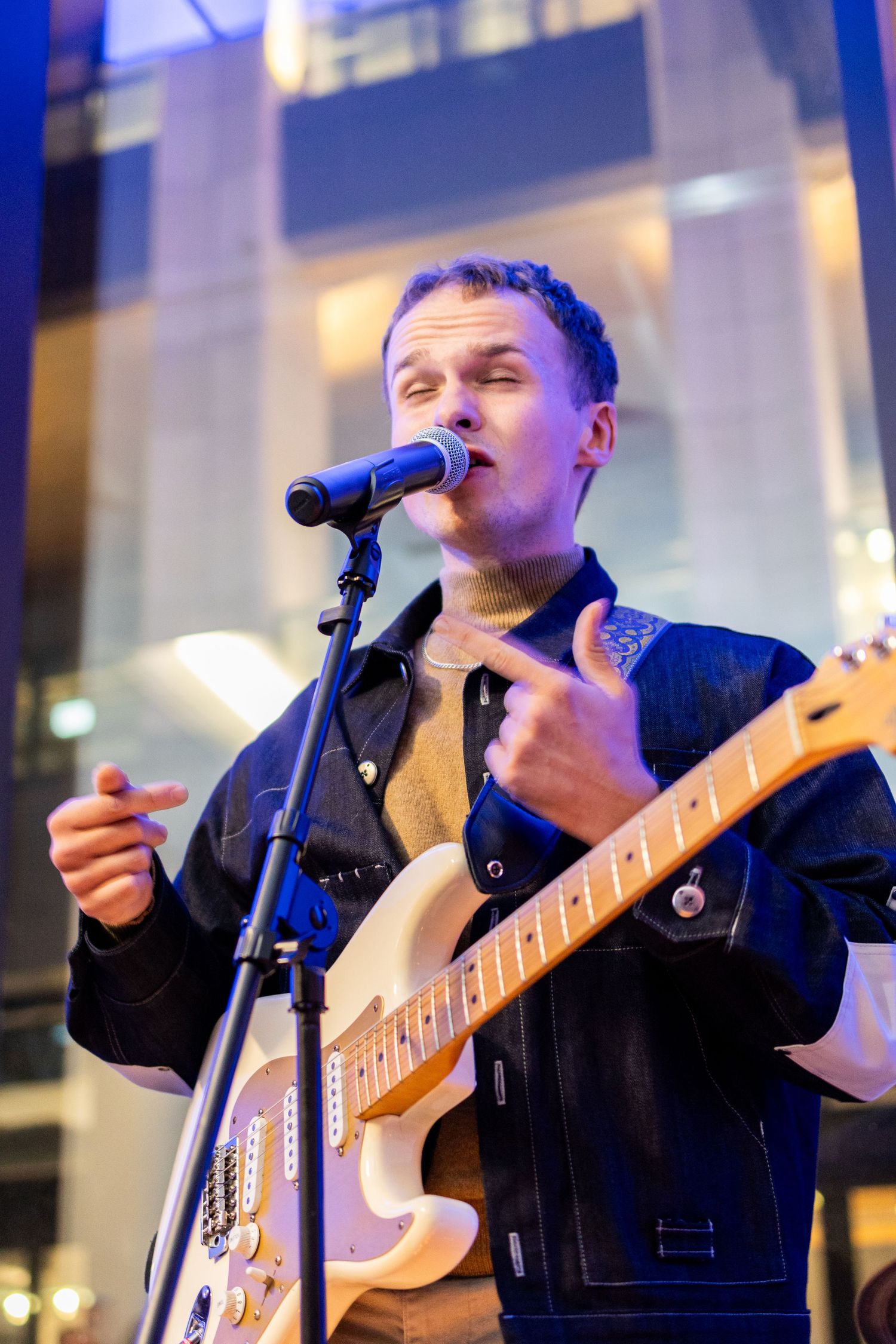 A musician in denim jacket performs while playing electric guitar on stage.