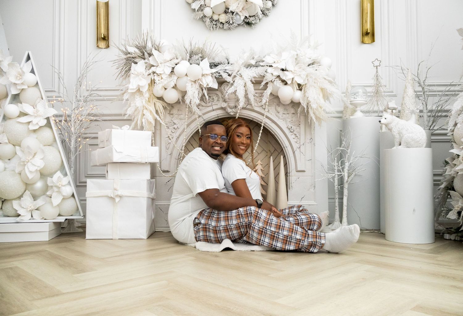 A loving couple embraces while sitting in front of an ornate white fireplace surrounded by festive holiday decorations.