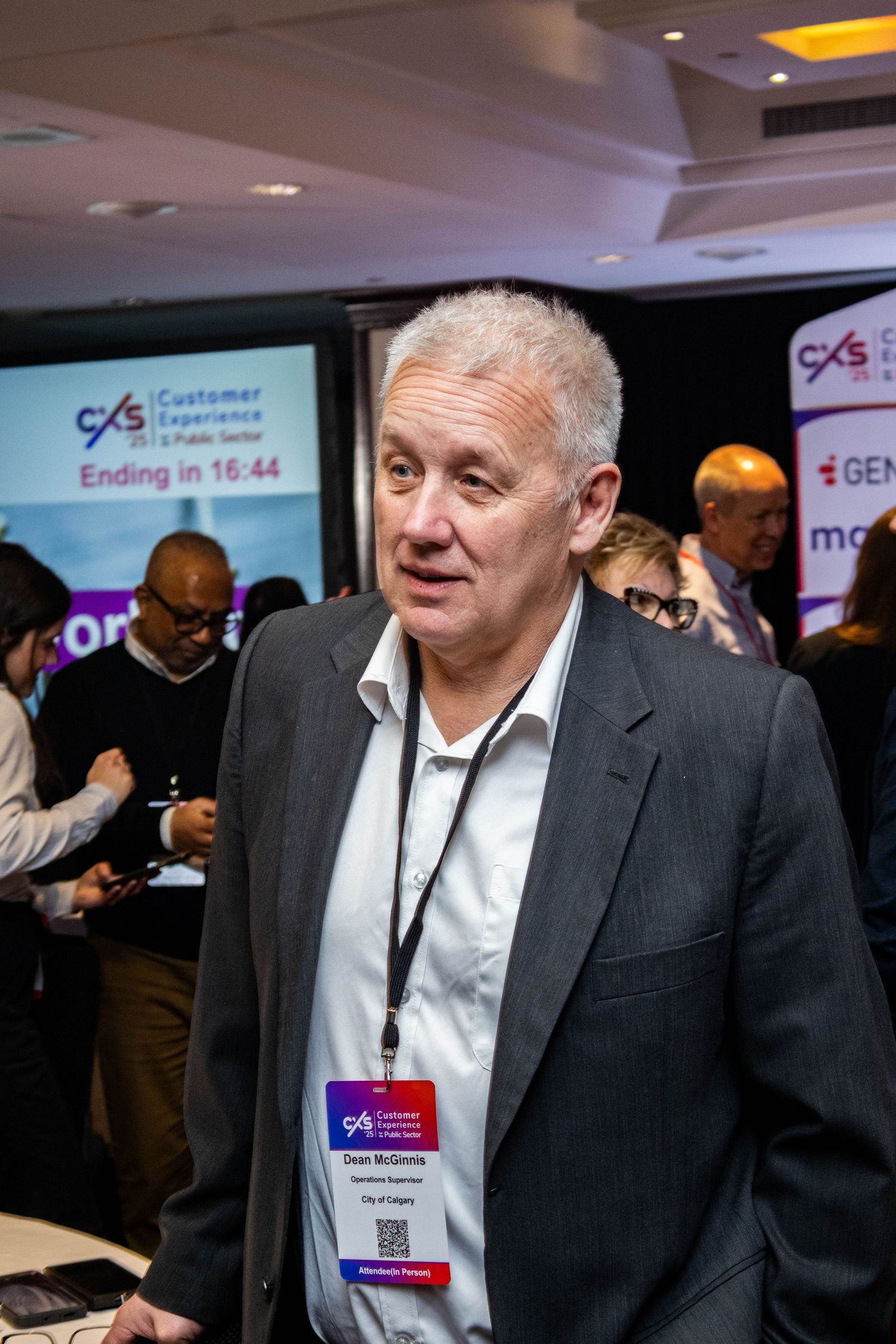 Business professional in grey suit jacket mingles at networking event with company banners visible.