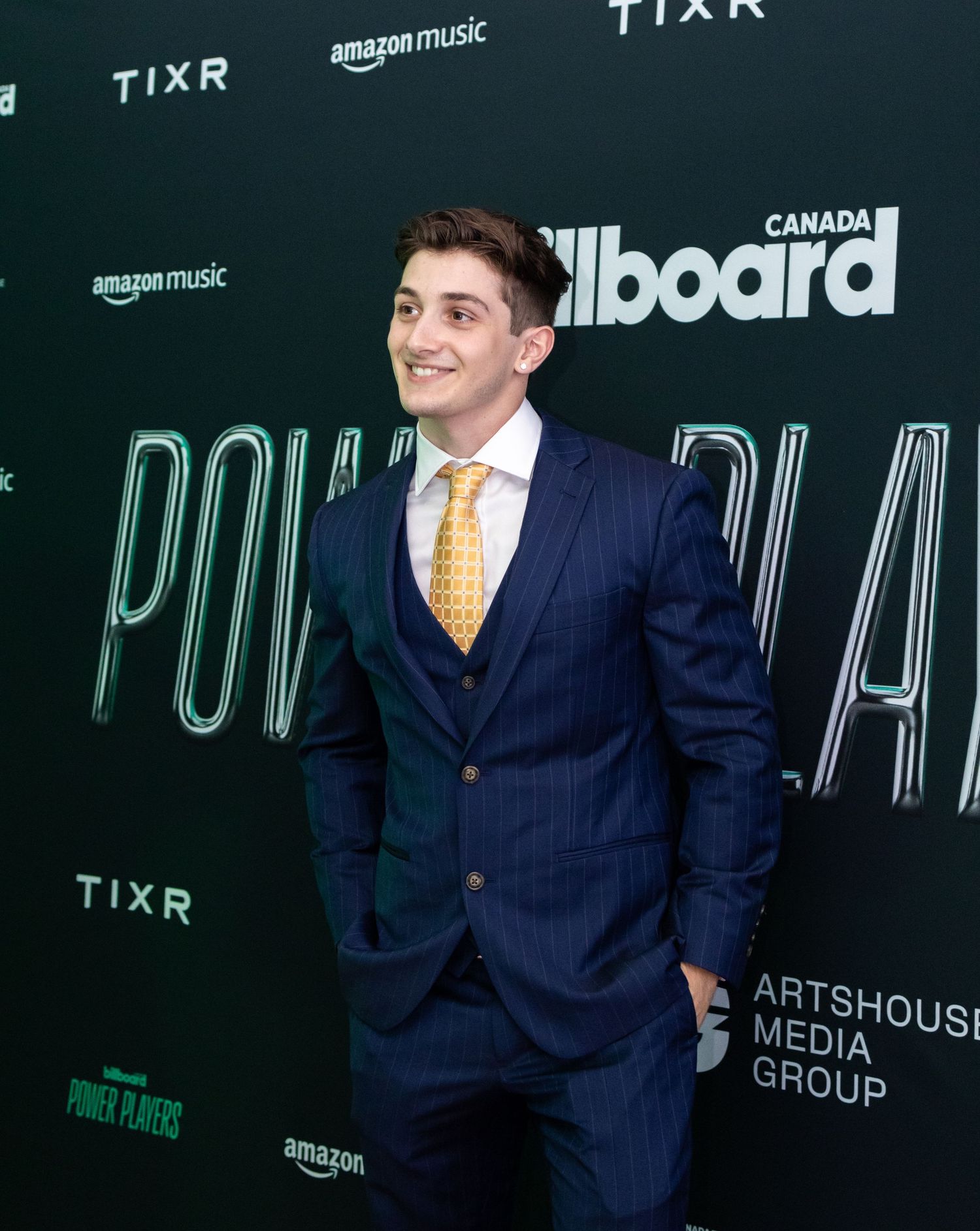 Young man in navy pinstripe suit with yellow tie poses at Billboard media event.