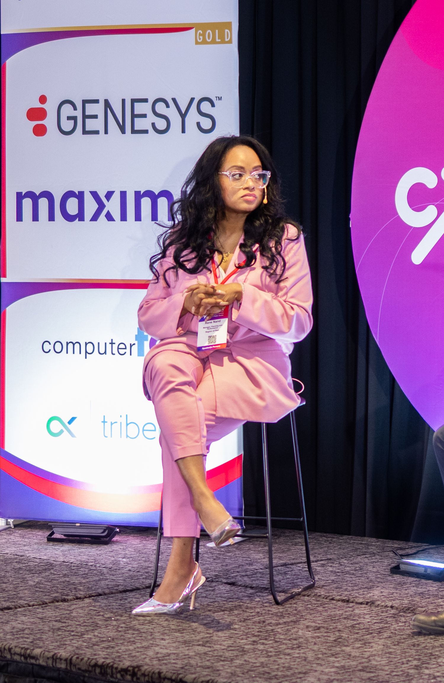 Conference speaker in pink pantsuit sits on stage with Genesys and Maximus company logos visible.