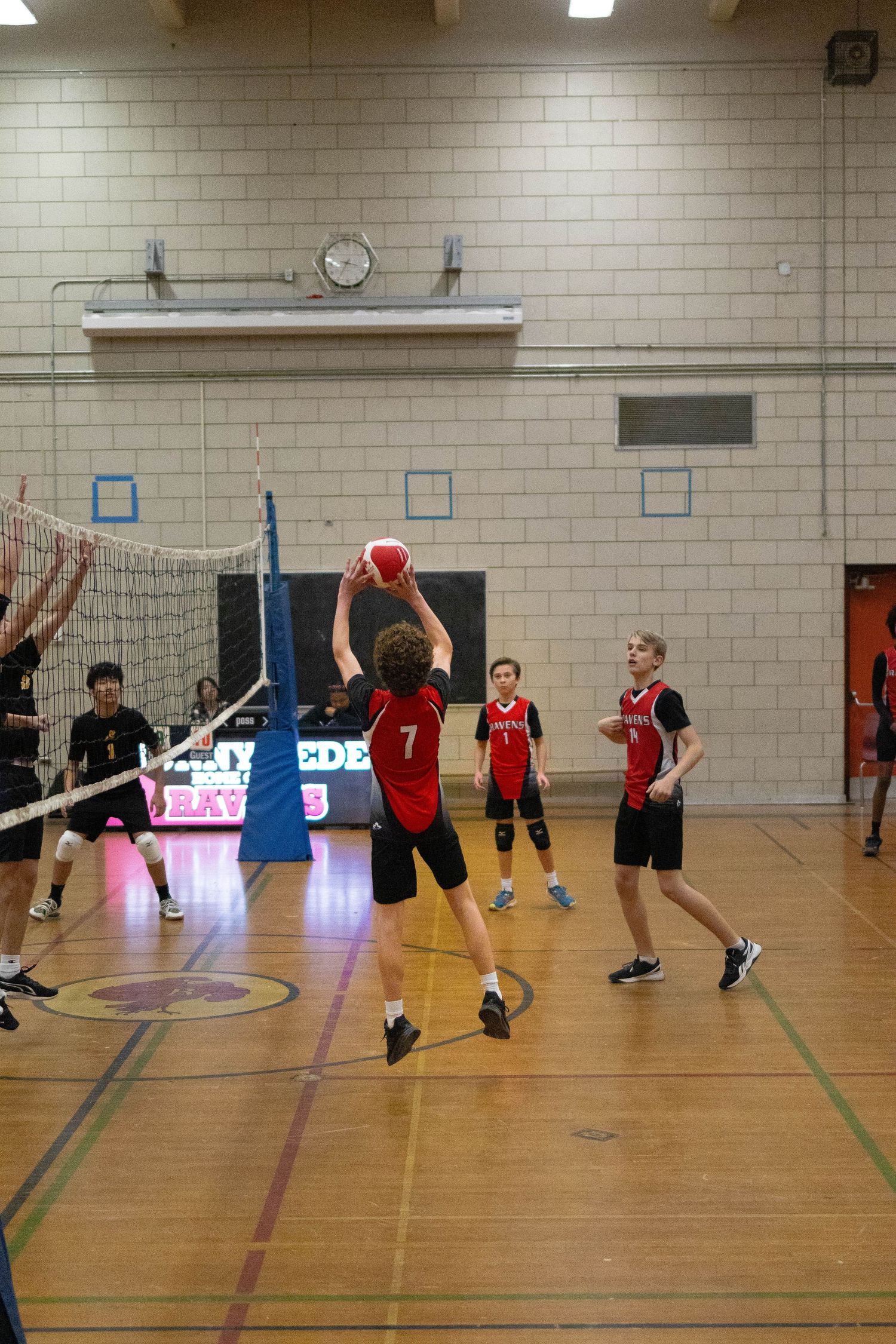Volleyball players competing in an indoor gymnasium with scoreboard visible in background.