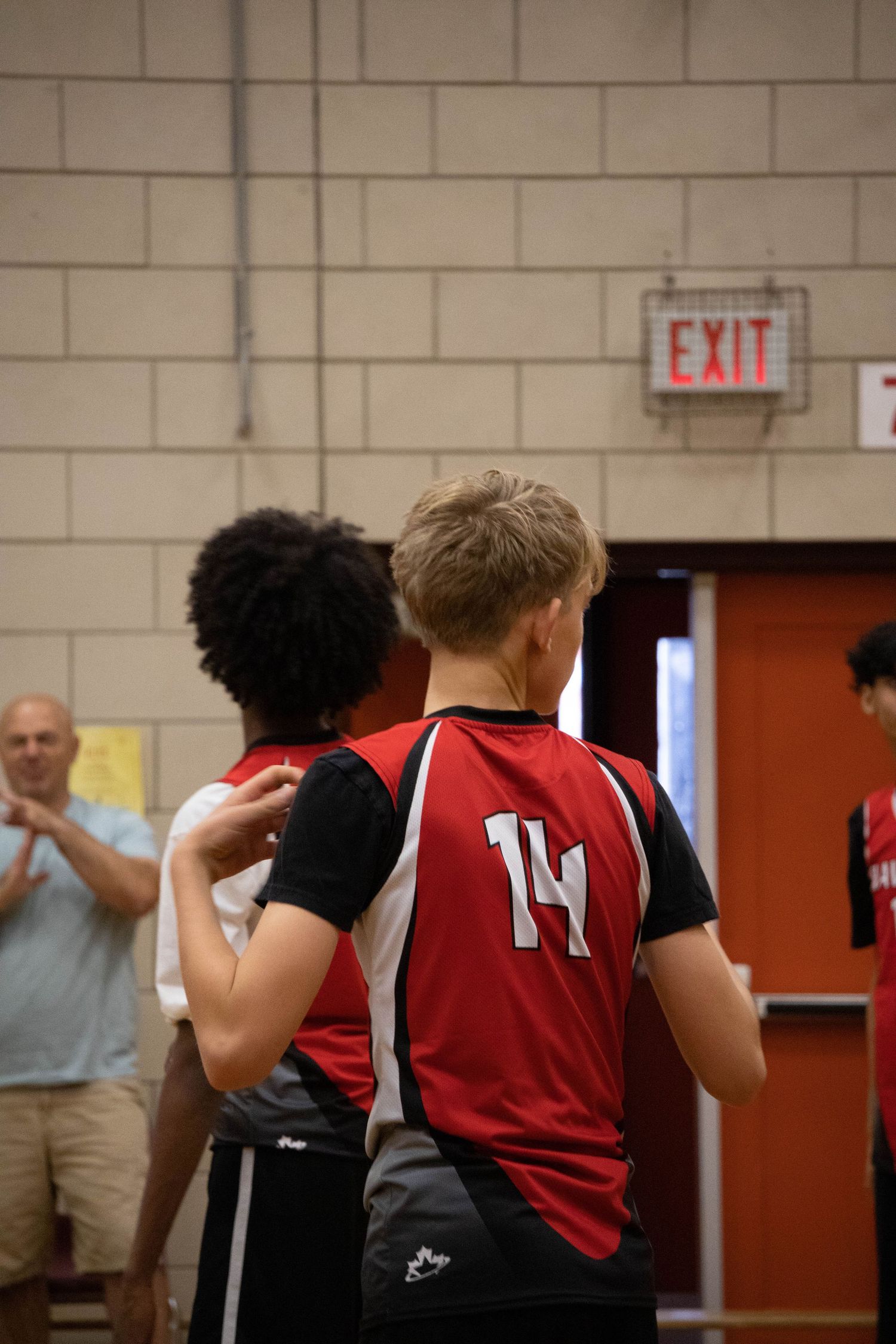 Two athletes in red and black jerseys standing near an exit sign in a gymnasium.