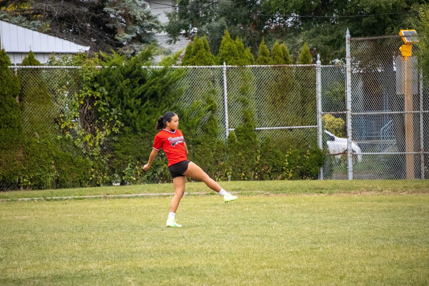 Player in red uniform kicking a ball on outdoor sports field with fence in background.