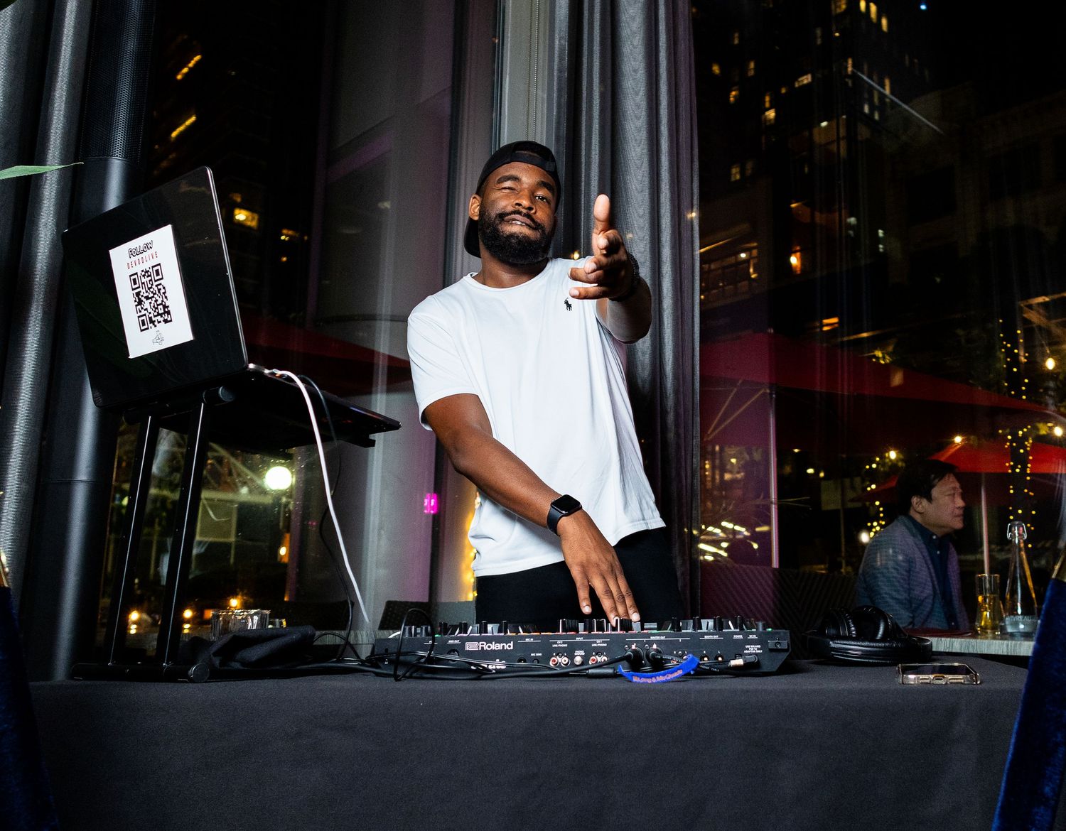 A DJ performs at an outdoor nighttime event with illuminated buildings visible in the background.