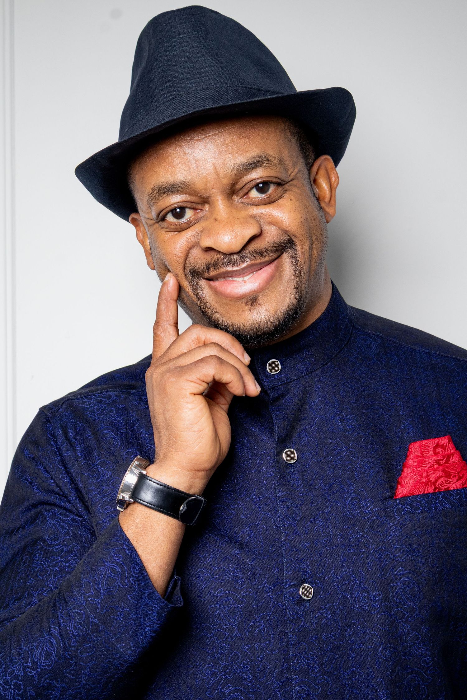 A portrait of someone in a navy fedora and mandarin collar jacket with red pocket square.
