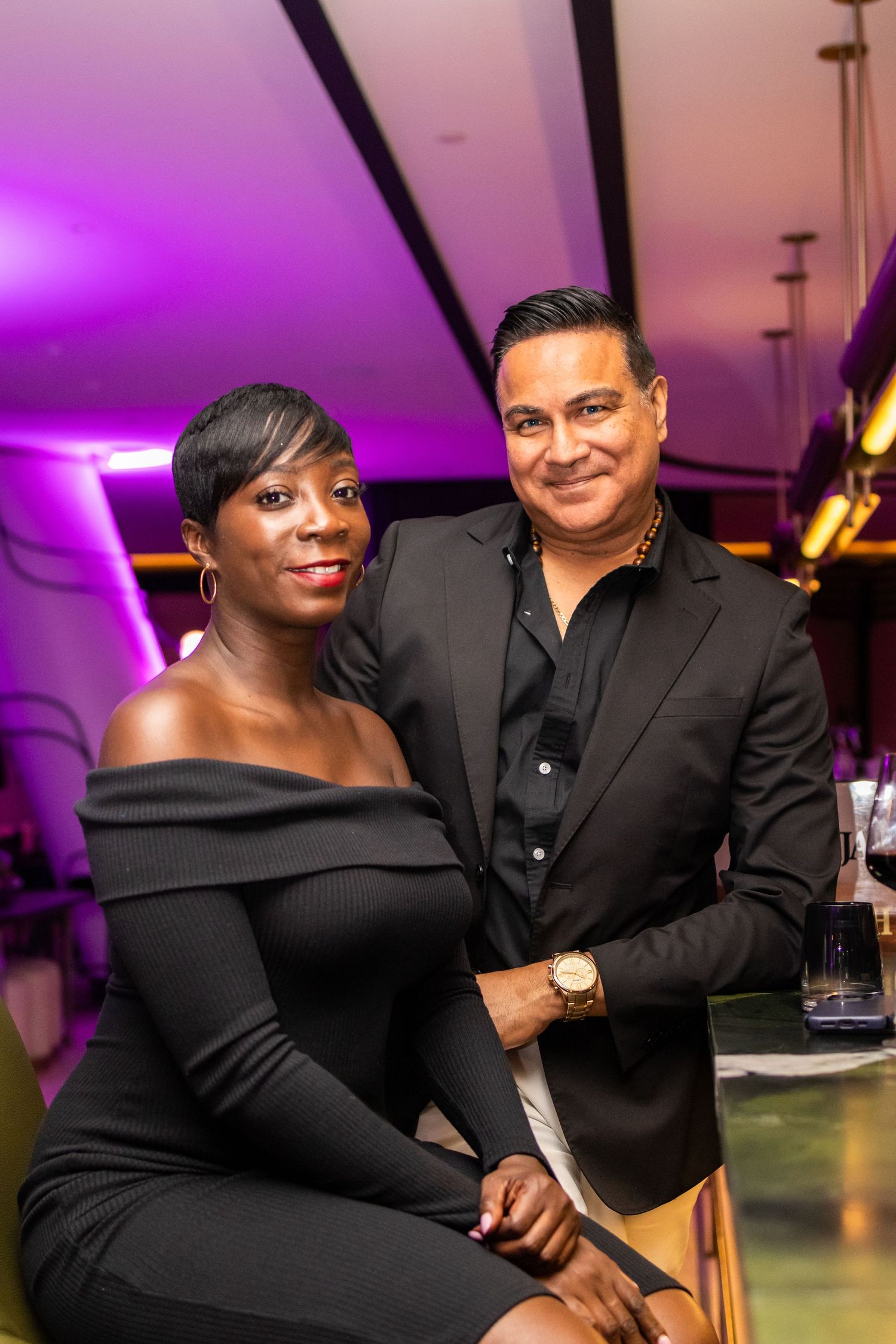 Elegant couple in formal black attire pose together at upscale venue with purple lighting.