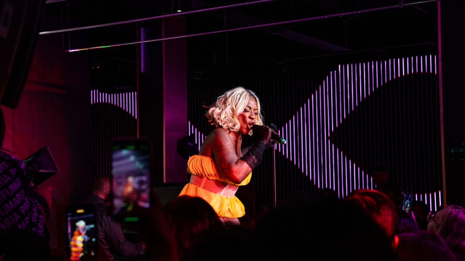 A performer in vibrant orange outfit sings on stage with dramatic purple lighting effects.