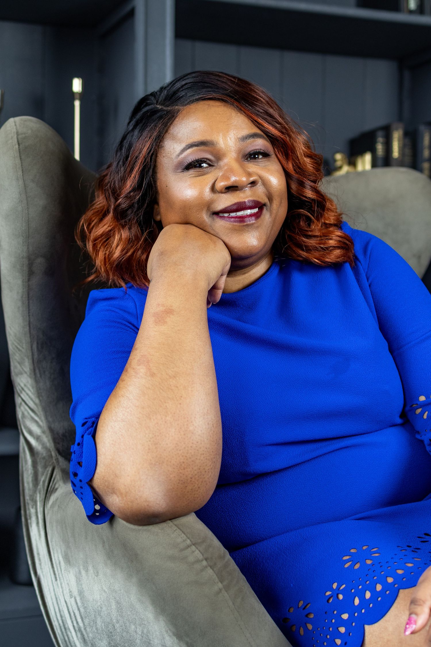 A person in a blue dress smiling while relaxing in a grey armchair.