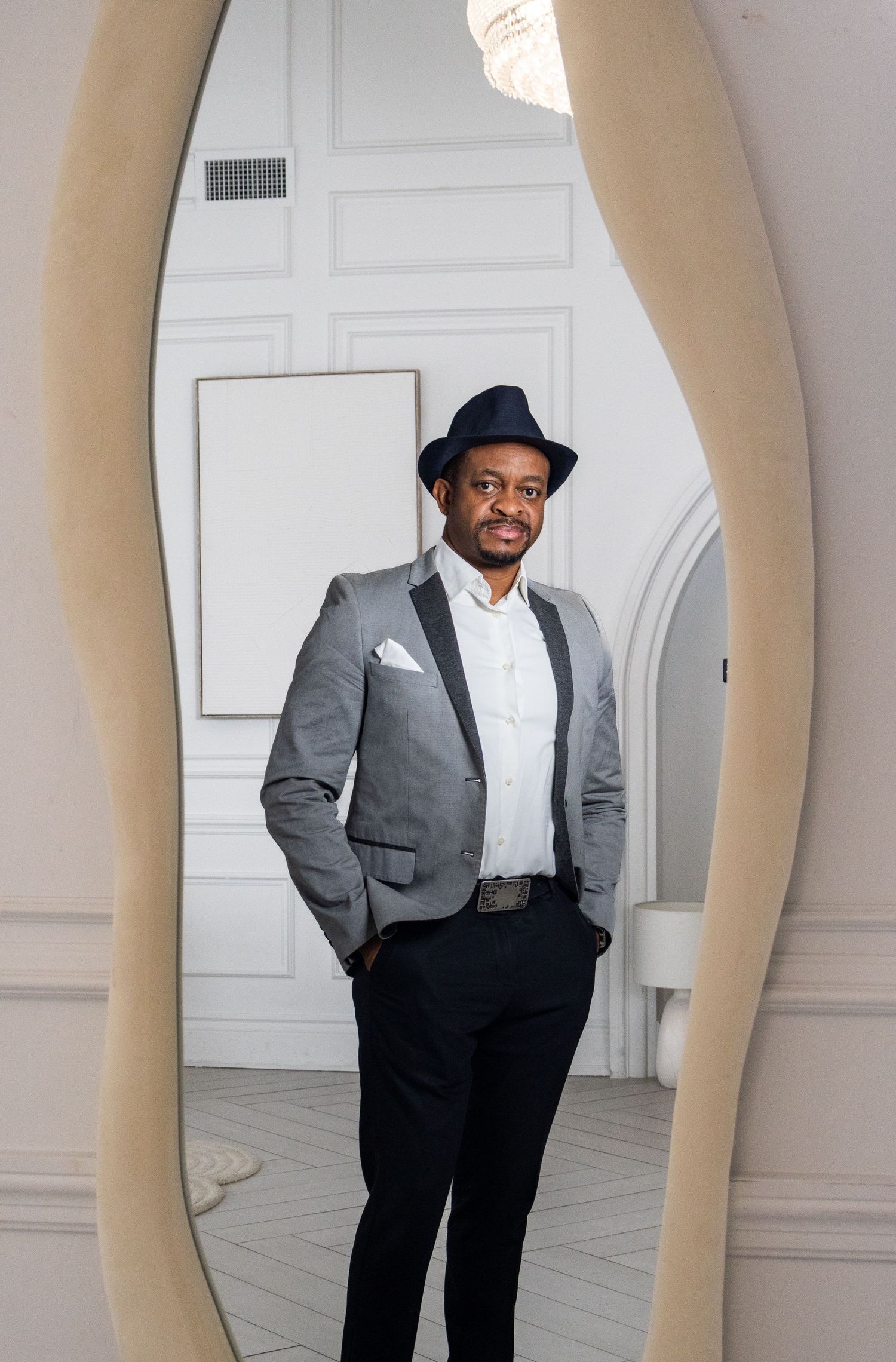 A stylish portrait through a mirror frame featuring someone in a grey suit and fedora.