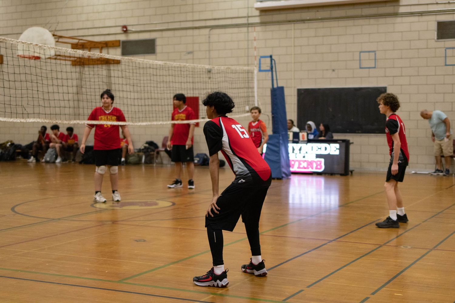 Indoor volleyball game with players in red uniforms positioned around the court during play.