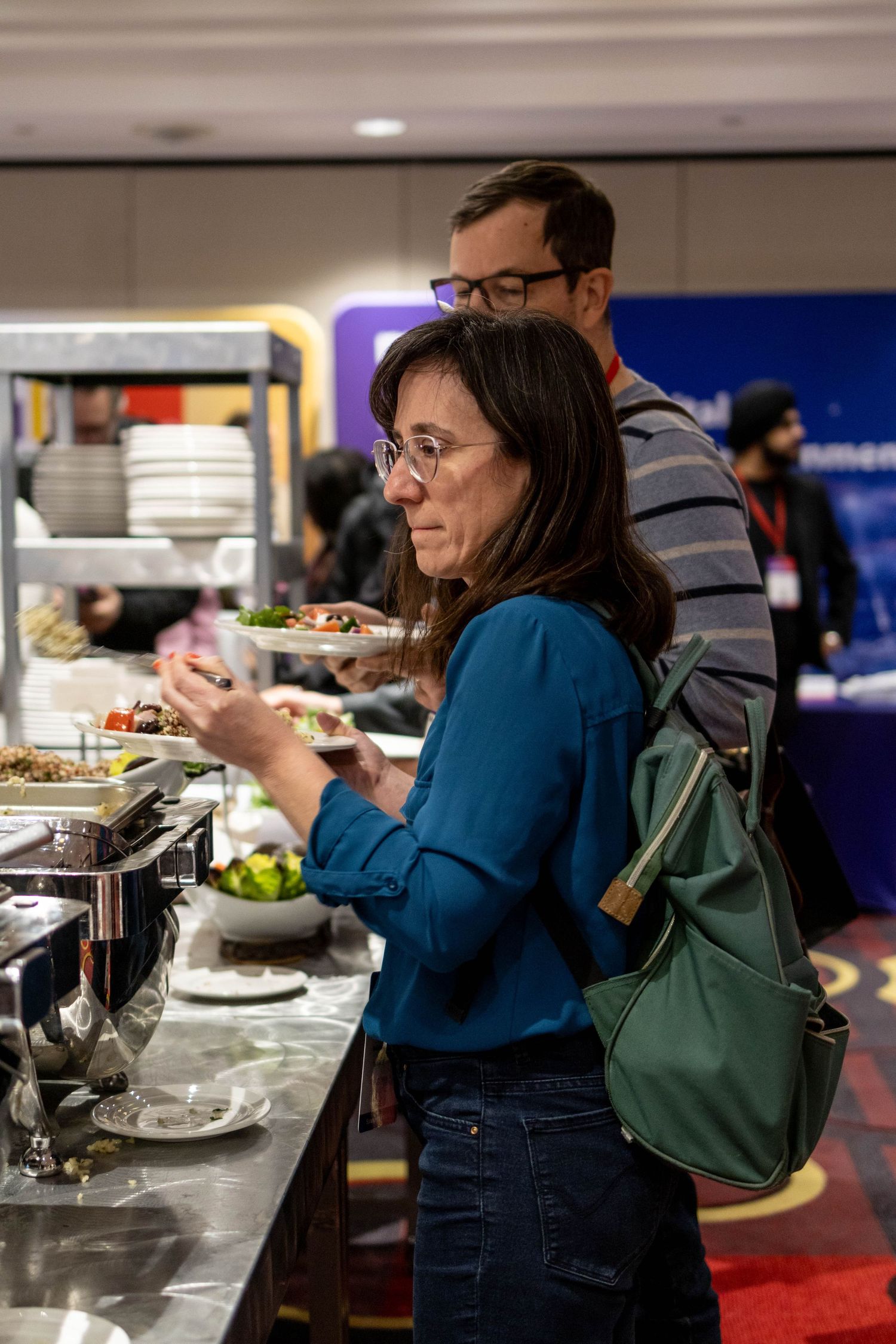Conference attendee in blue sweater selects food from a buffet line while wearing a green shoulder bag.