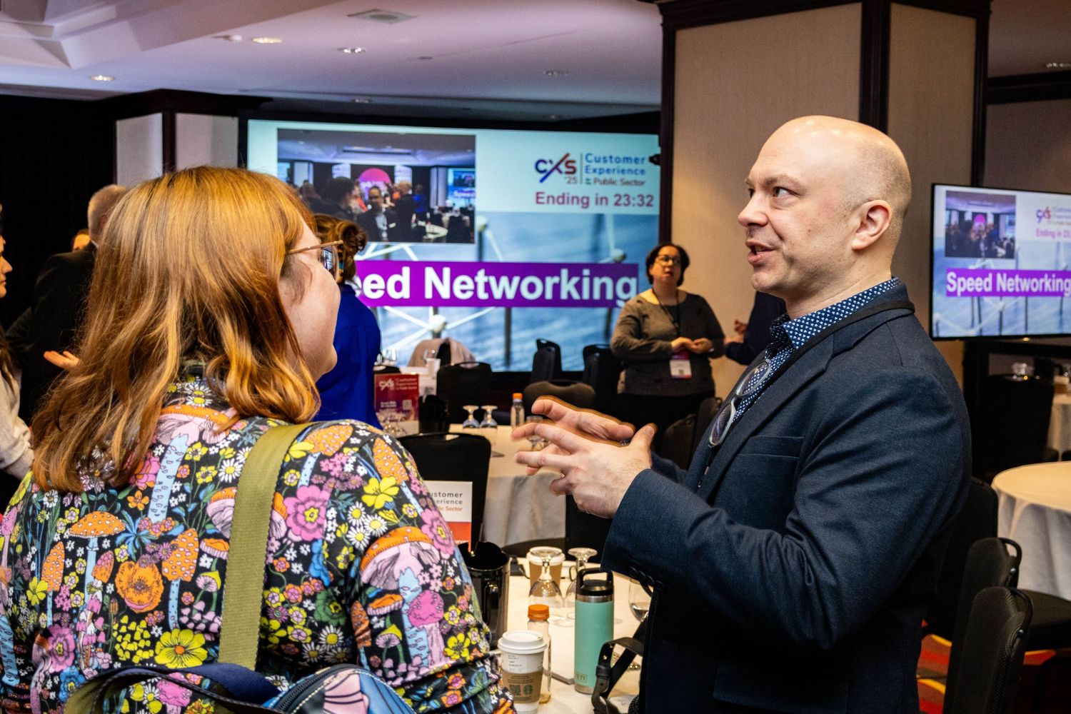 Two professionals engaged in animated discussion at networking event with screens visible in background.