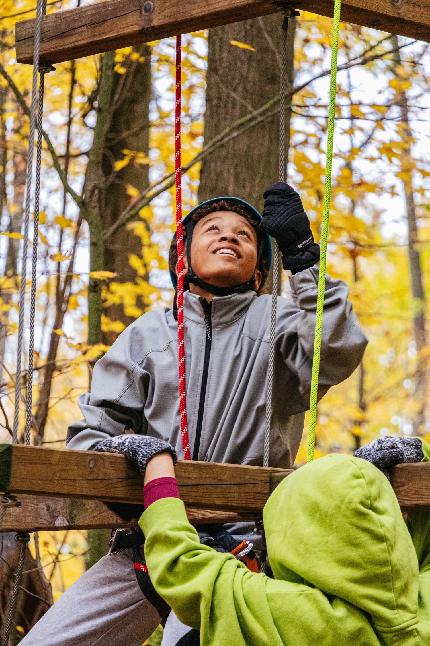 Young adventurer climbs on ropes course wearing safety gear amid autumn foliage.
