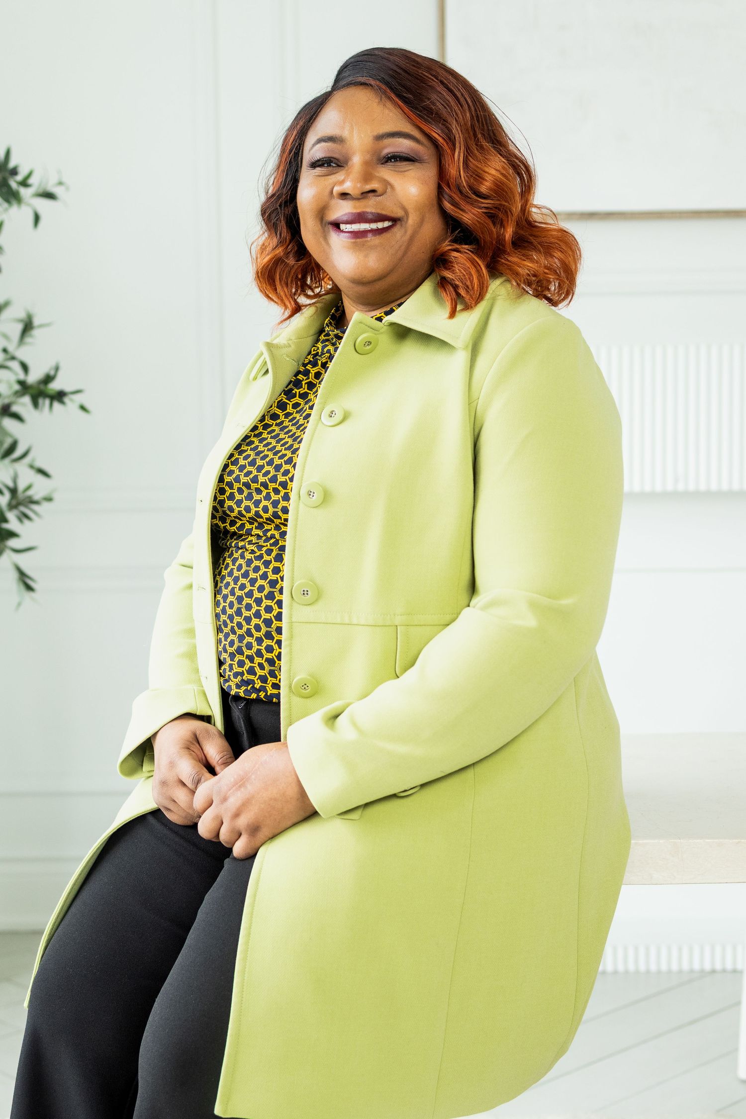Stylish woman in bright yellow coat smiles warmly while seated in contemporary white room.